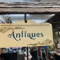 Beige “Antiques” shop sign with decorative brown scrollwork hanging outside a storefront