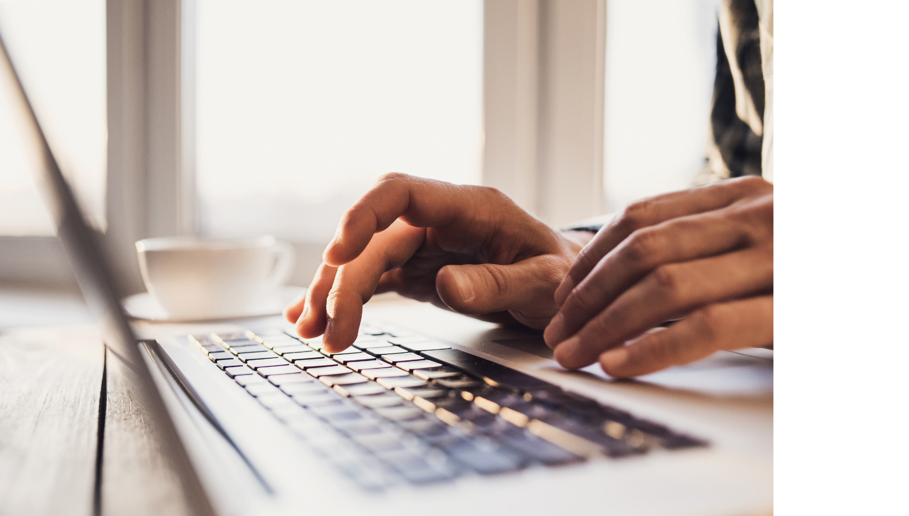 Hands typing on a laptop keyboard at a desk next to a white coffee cup in a brightly lit room.