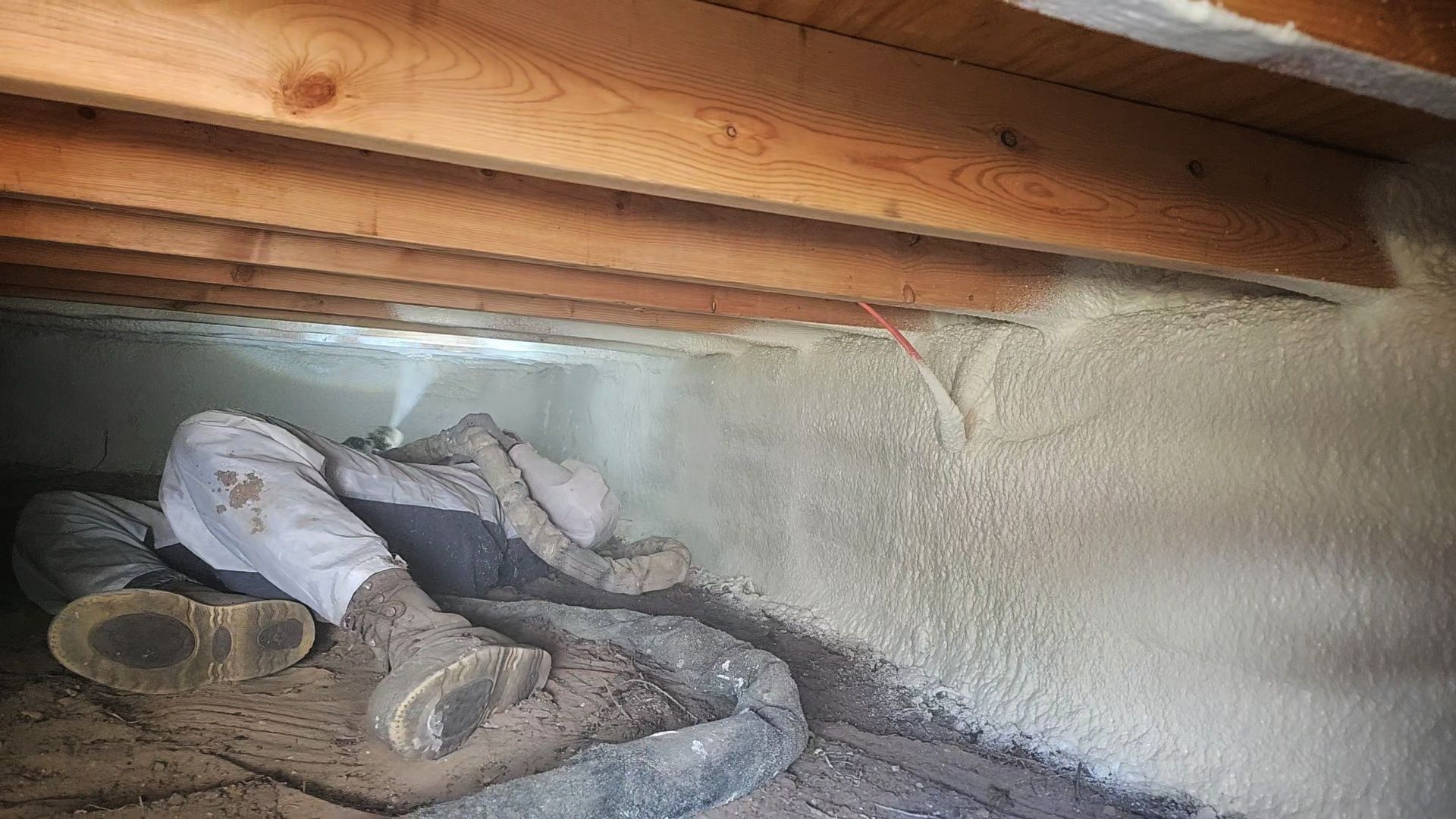 Man in protective gear spraying insulation under wooden beams.