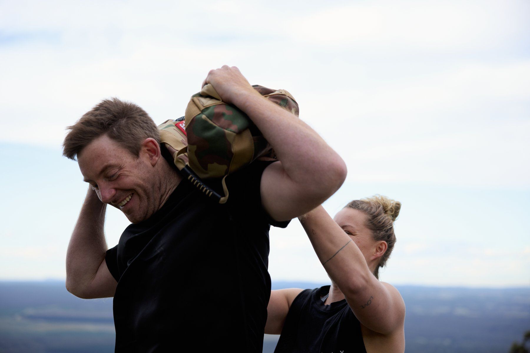 Two hikers carrying large backpacks uphill, one grimacing, with a cloudy landscape behind them