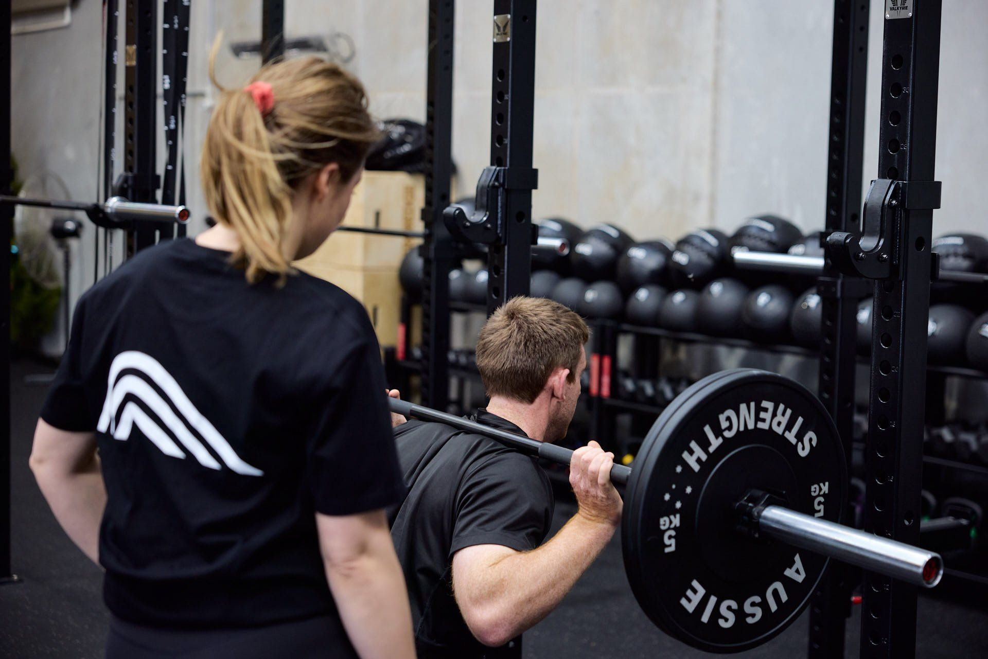 Woman spotting a man back squatting a barbell in a gym, with weight plates and kettlebells behind them