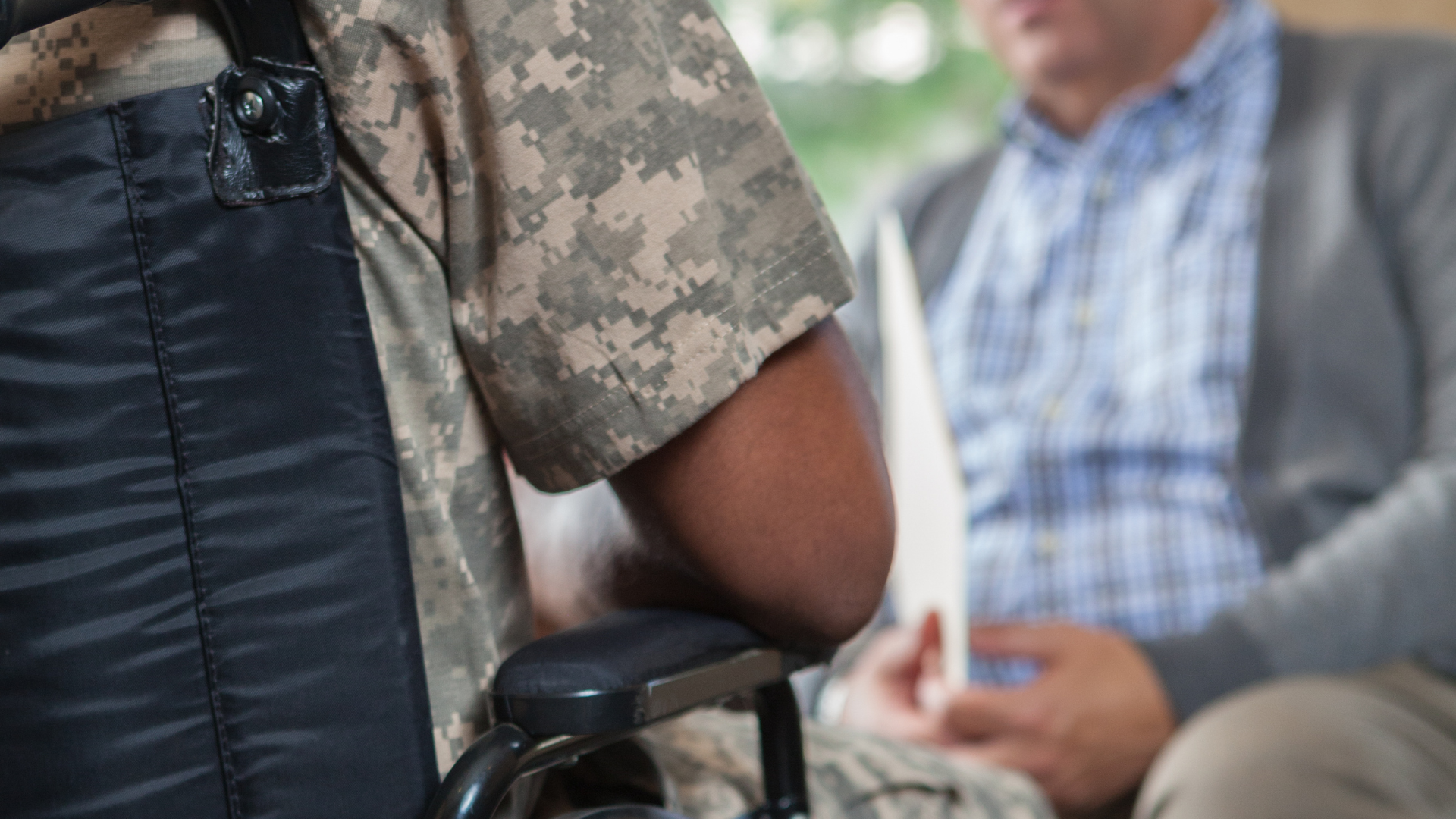 Soldier in camouflage uniform sitting in a wheelchair, facing an older man in a light-colored sweater.