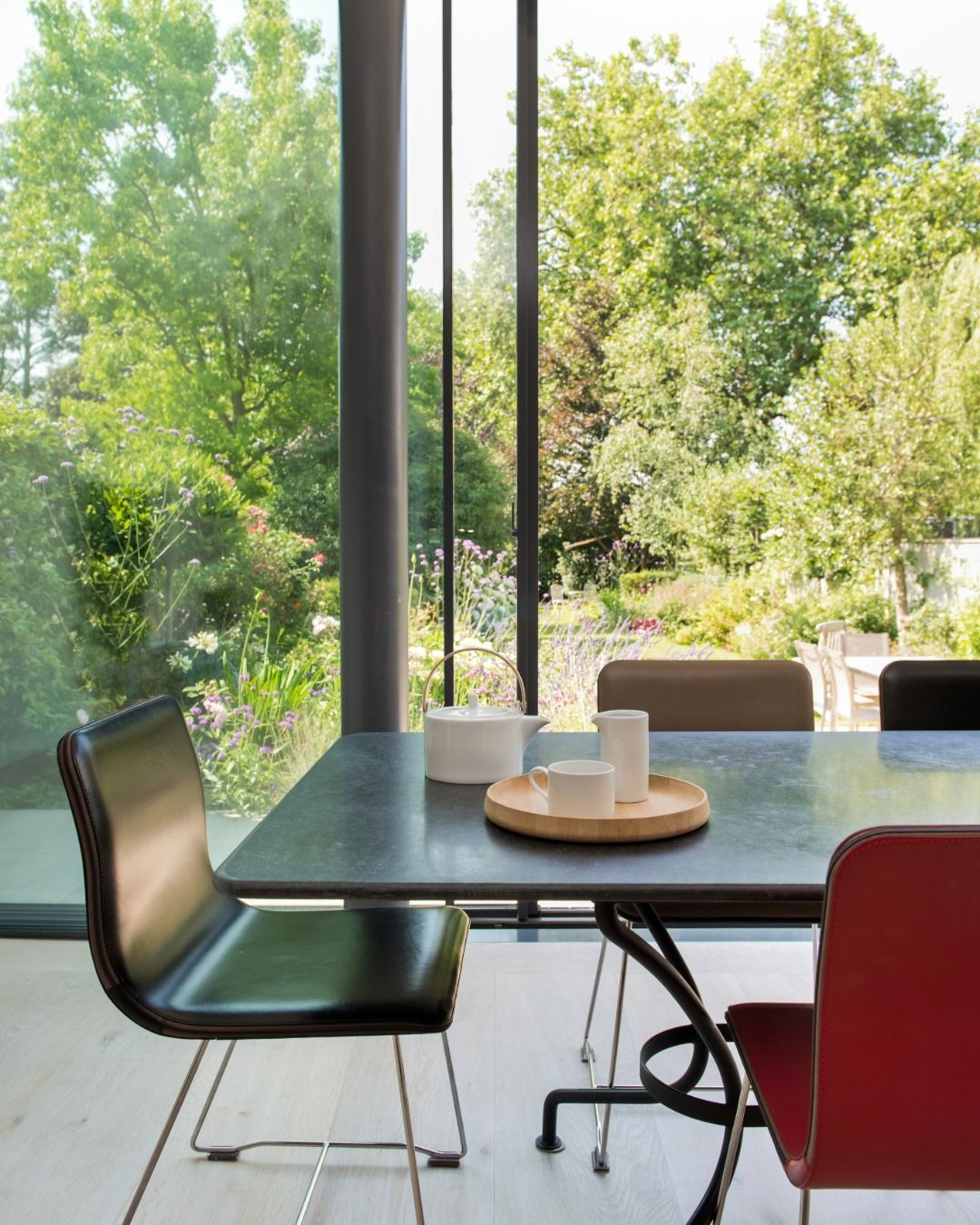 Modern dining table with mixed chairs beside large windows overlooking a green garden.