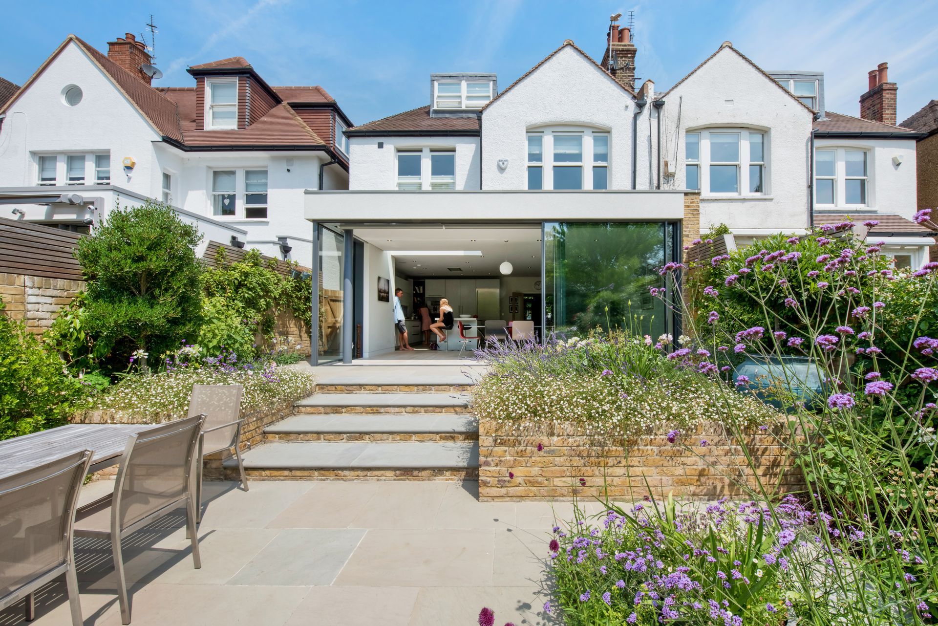 Backyard patio with steps leading to a modern house and open glass doors, bordered by garden plants.