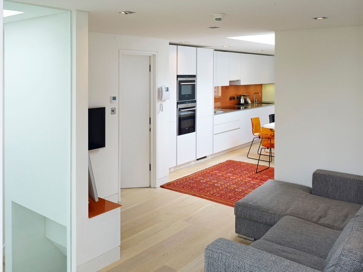 Modern open-plan kitchen and living room with white cabinets, orange backsplash, and gray sofa on light wood floor