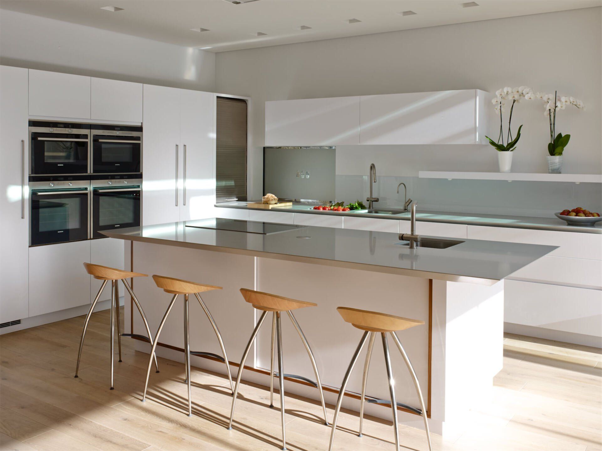 Modern white kitchen with island, bar stools, built-in ovens, and orchids on the counter