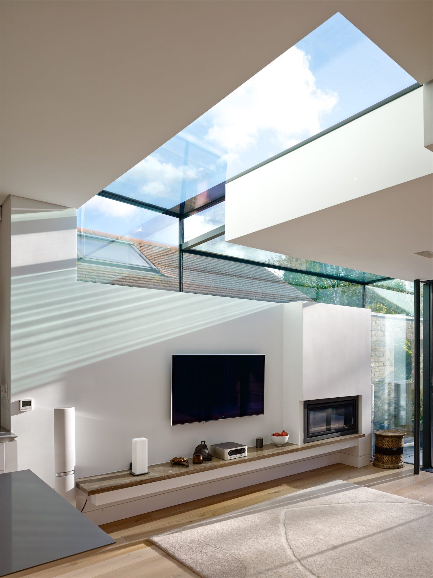 Modern living room with tall glass windows, skylight, wall-mounted TV, fireplace, and a long wooden console