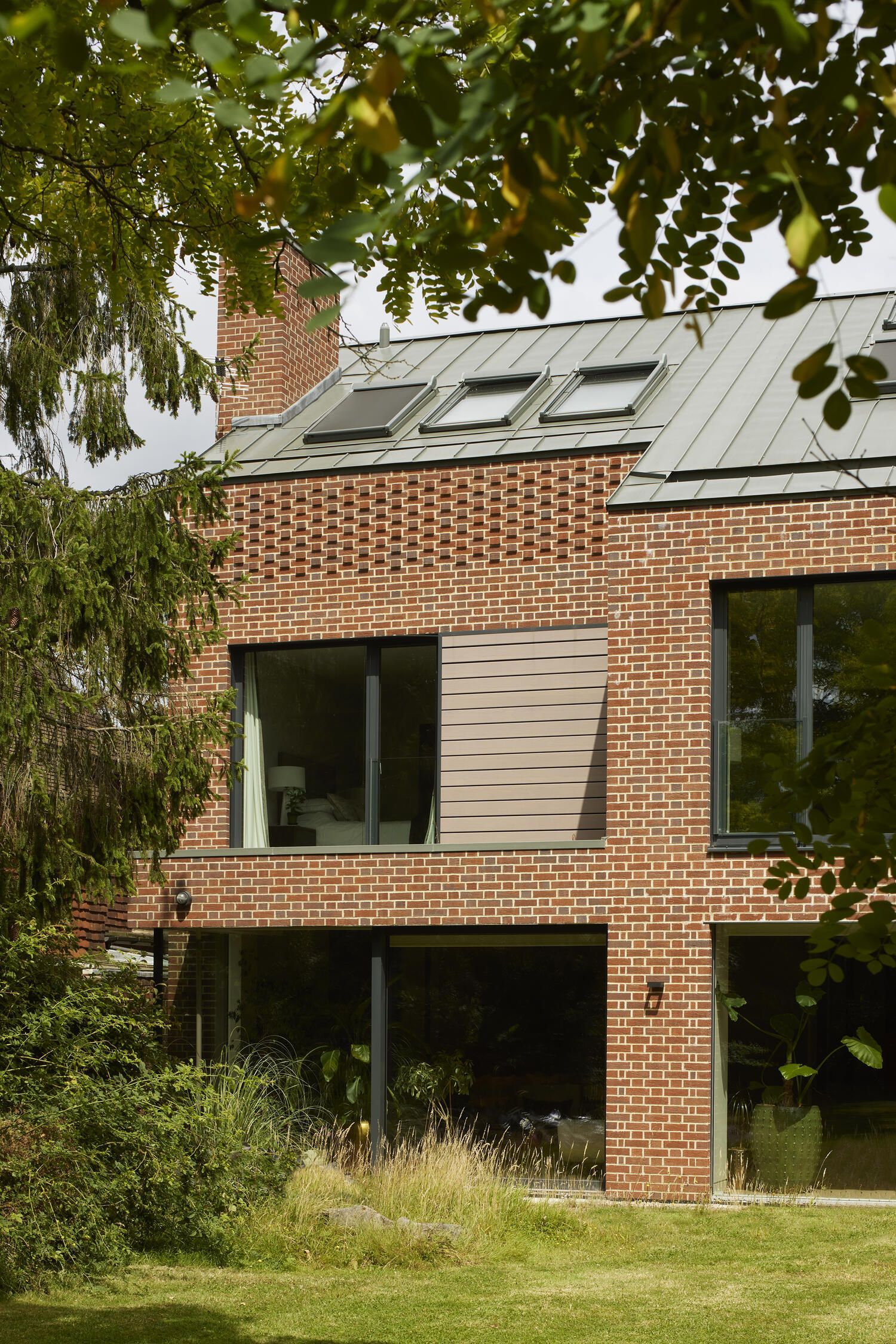 Modern brick house with large windows and a sloped roof, partially obscured by leafy trees and greenery.