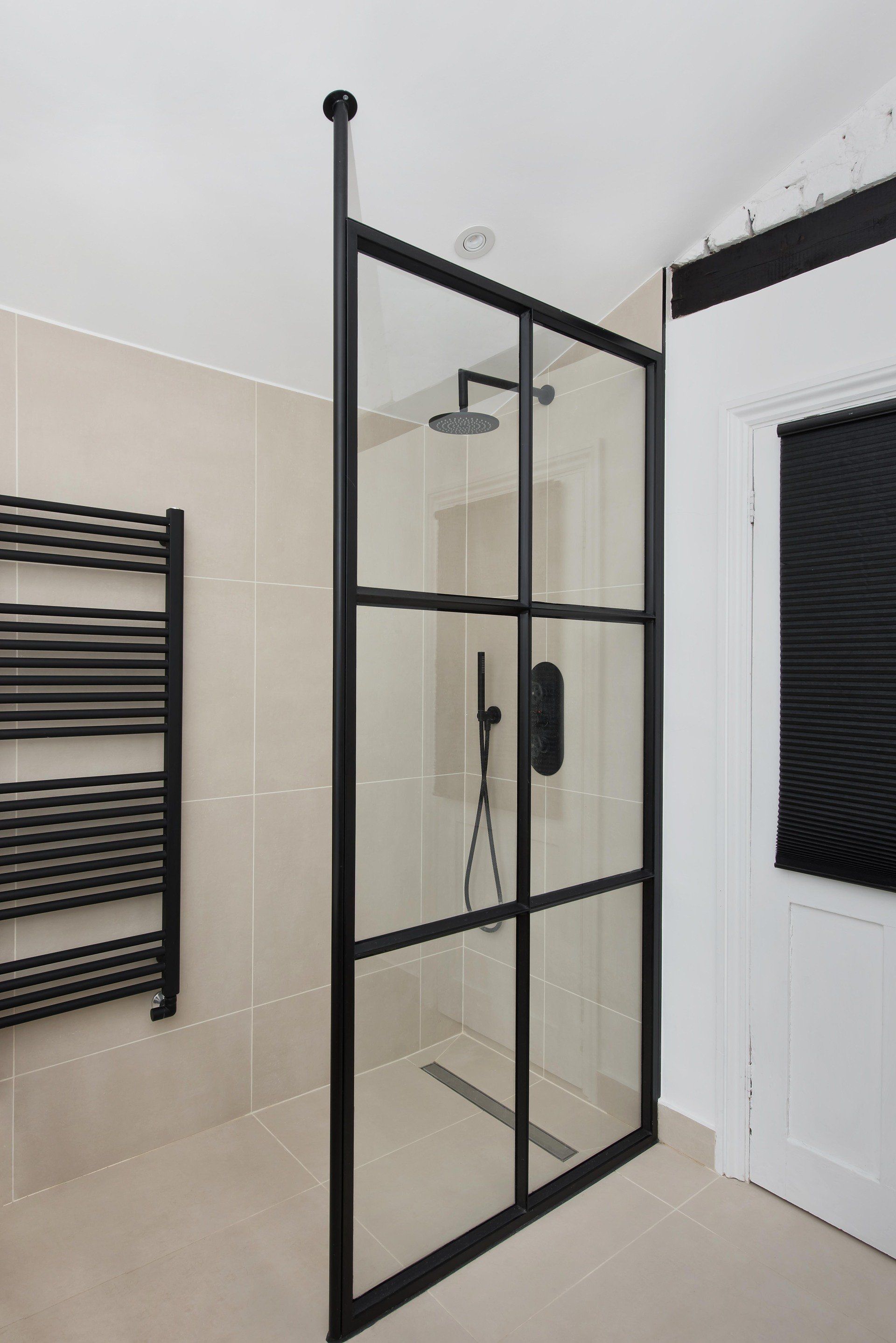Black-framed glass shower enclosure in a beige-tiled bathroom beside a white door.