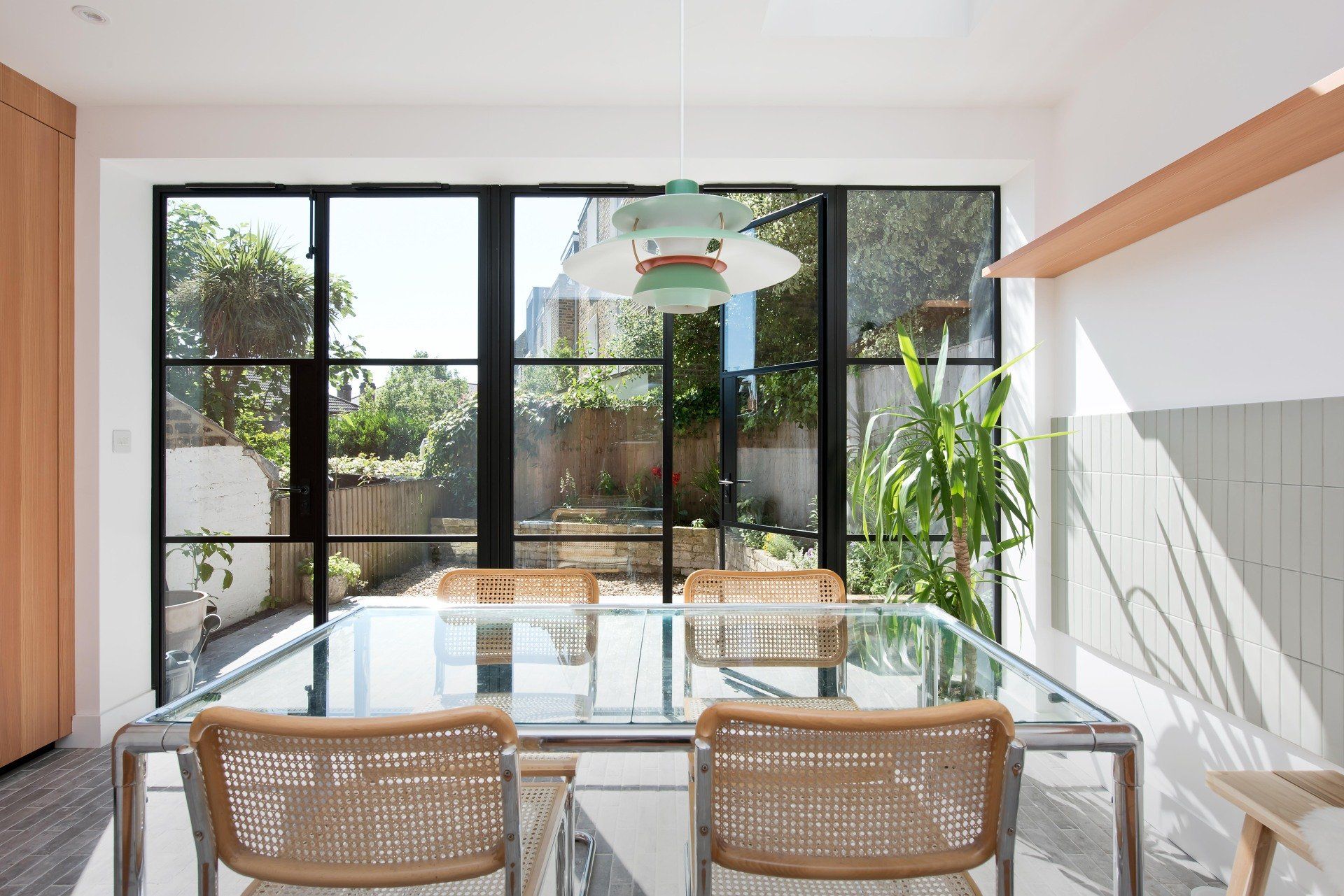 Bright dining room with glass table, woven chairs, and large windows opening to a leafy garden.