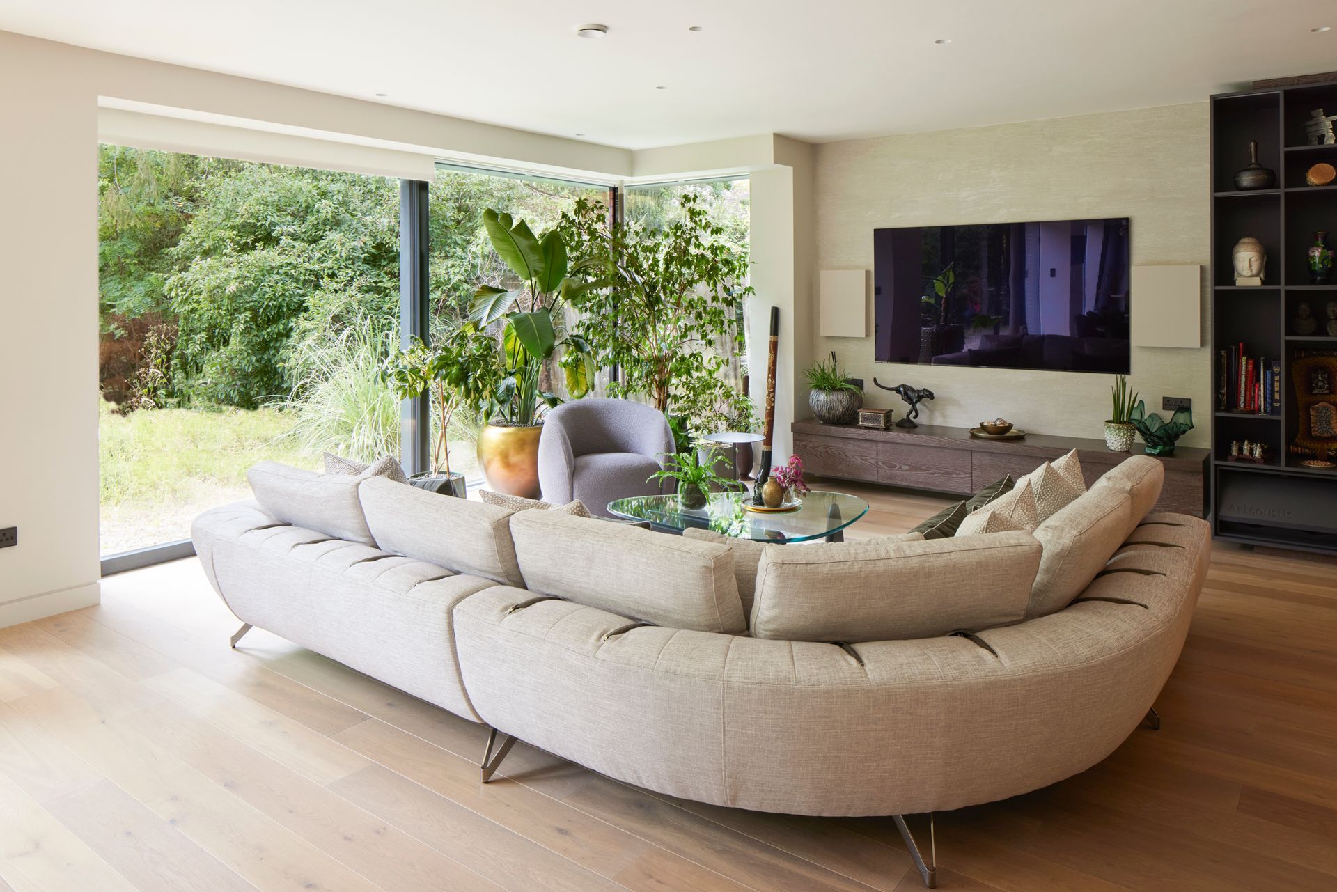 Modern living room with a curved beige sectional, wall-mounted TV, and open view to a green garden