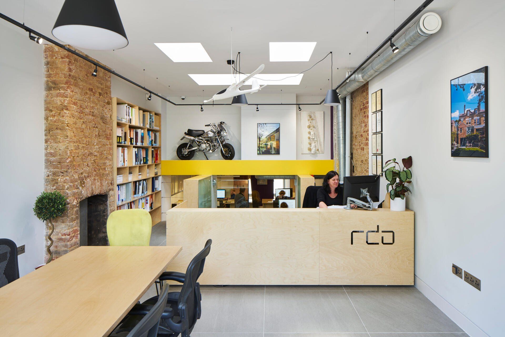 Modern office reception area with light wood desks, bookshelves, yellow accent wall, and a person behind the counter