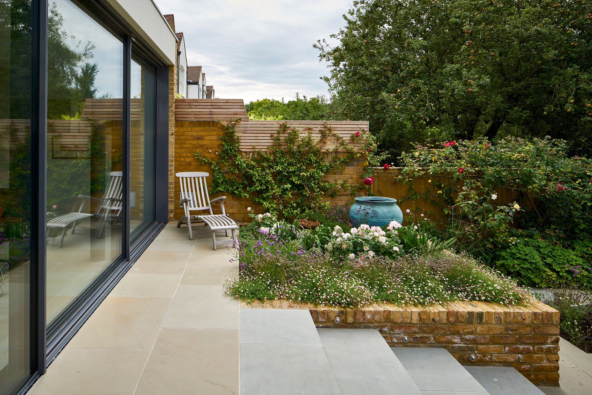 Patio with glass doors, chairs, flower beds, and a green garden under a cloudy sky