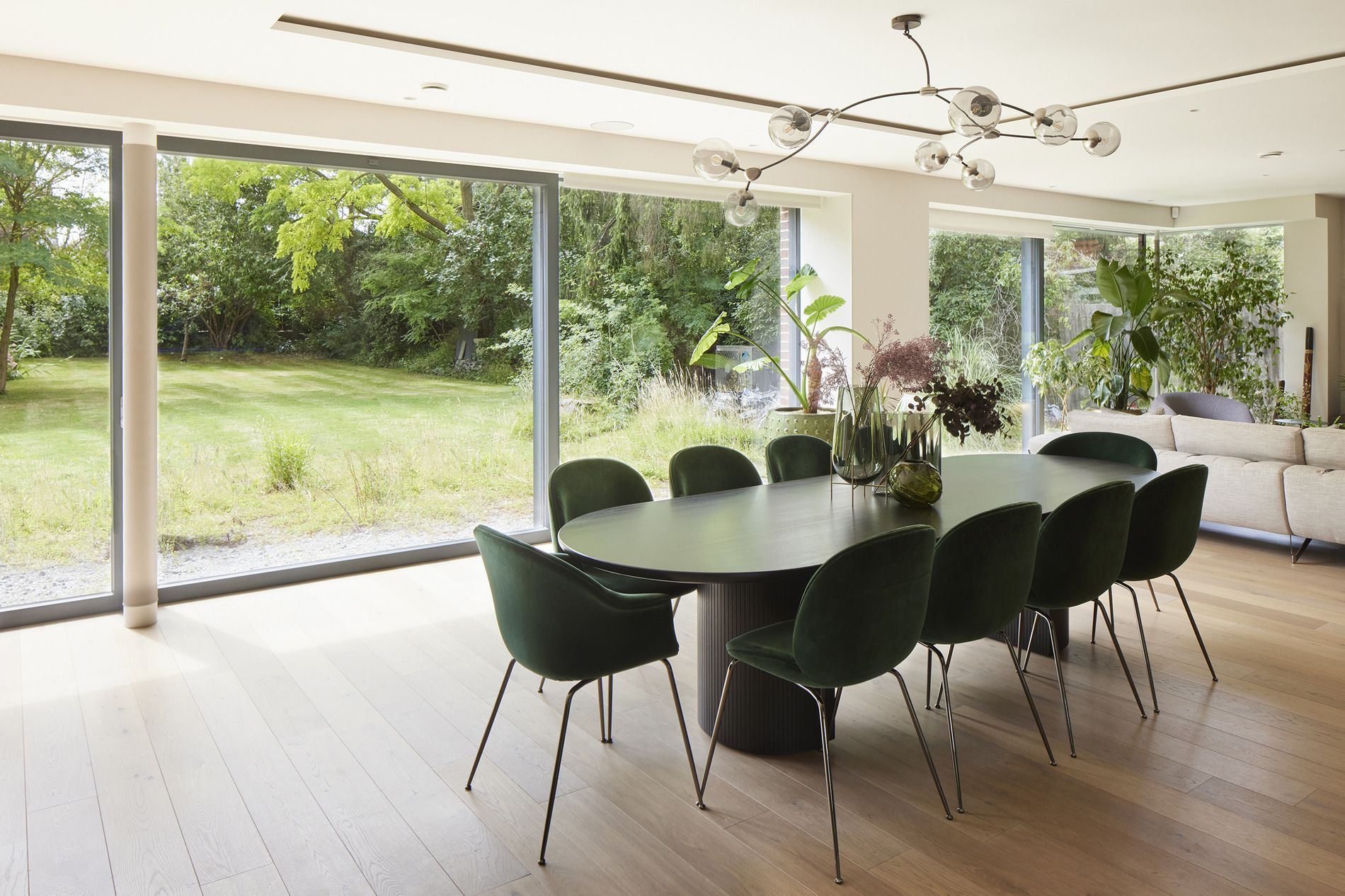 Modern dining room with black chairs and a wooden table beside floor-to-ceiling windows overlooking a green garden