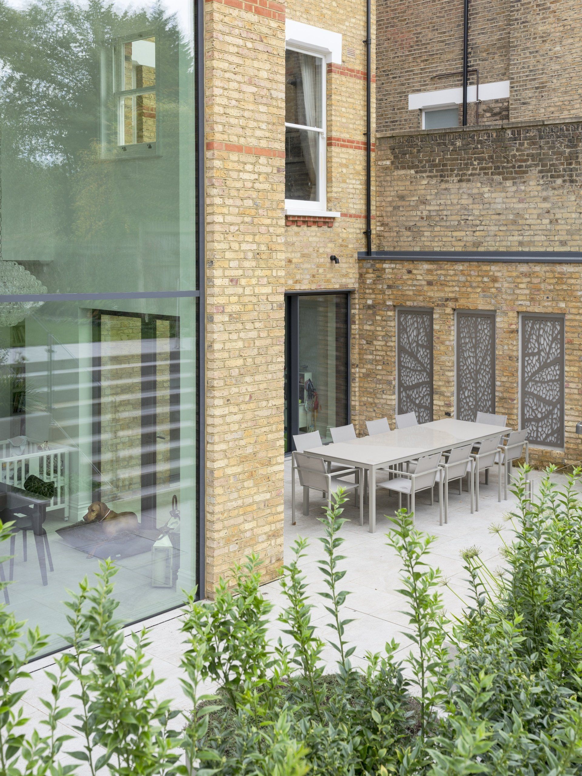 Patio with long outdoor dining table beside brick and glass building, framed by greenery