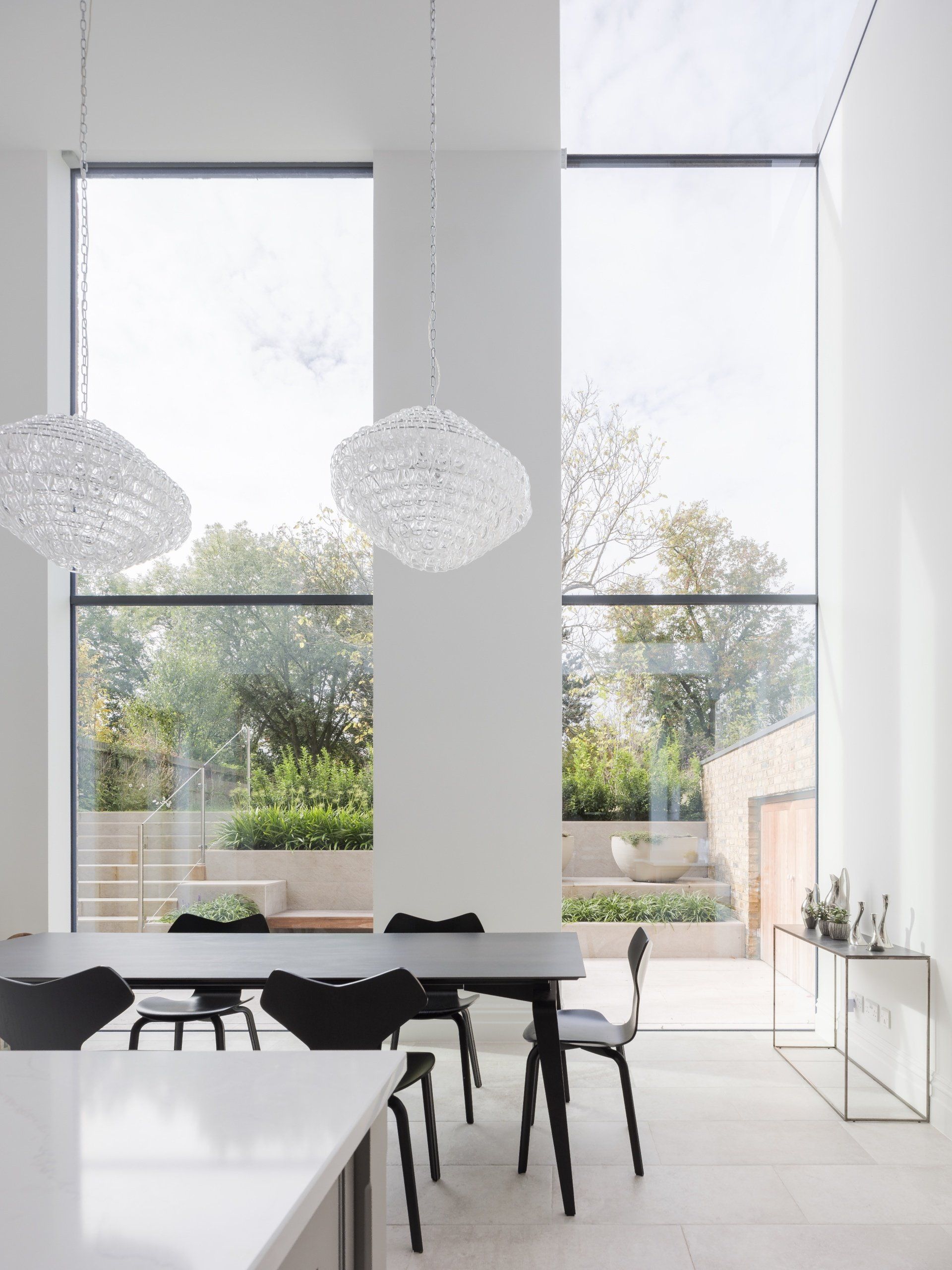 Modern minimalist dining room with tall windows, white walls, black chairs, and pendant lights overlooking greenery