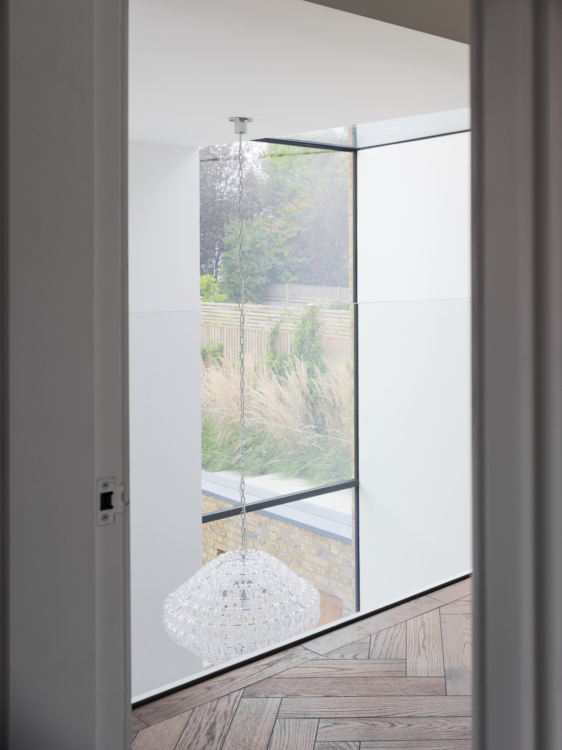 Modern entryway with glass doors, white walls, and herringbone wood flooring, opening to a leafy outdoor view