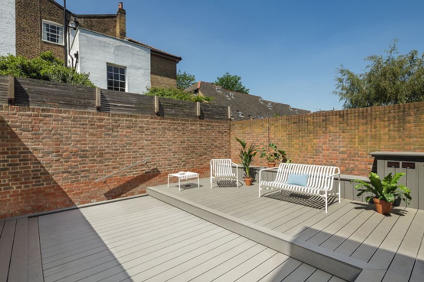 Brick patio with light gray decking, white bench, potted plants, and brick walls under a blue sky