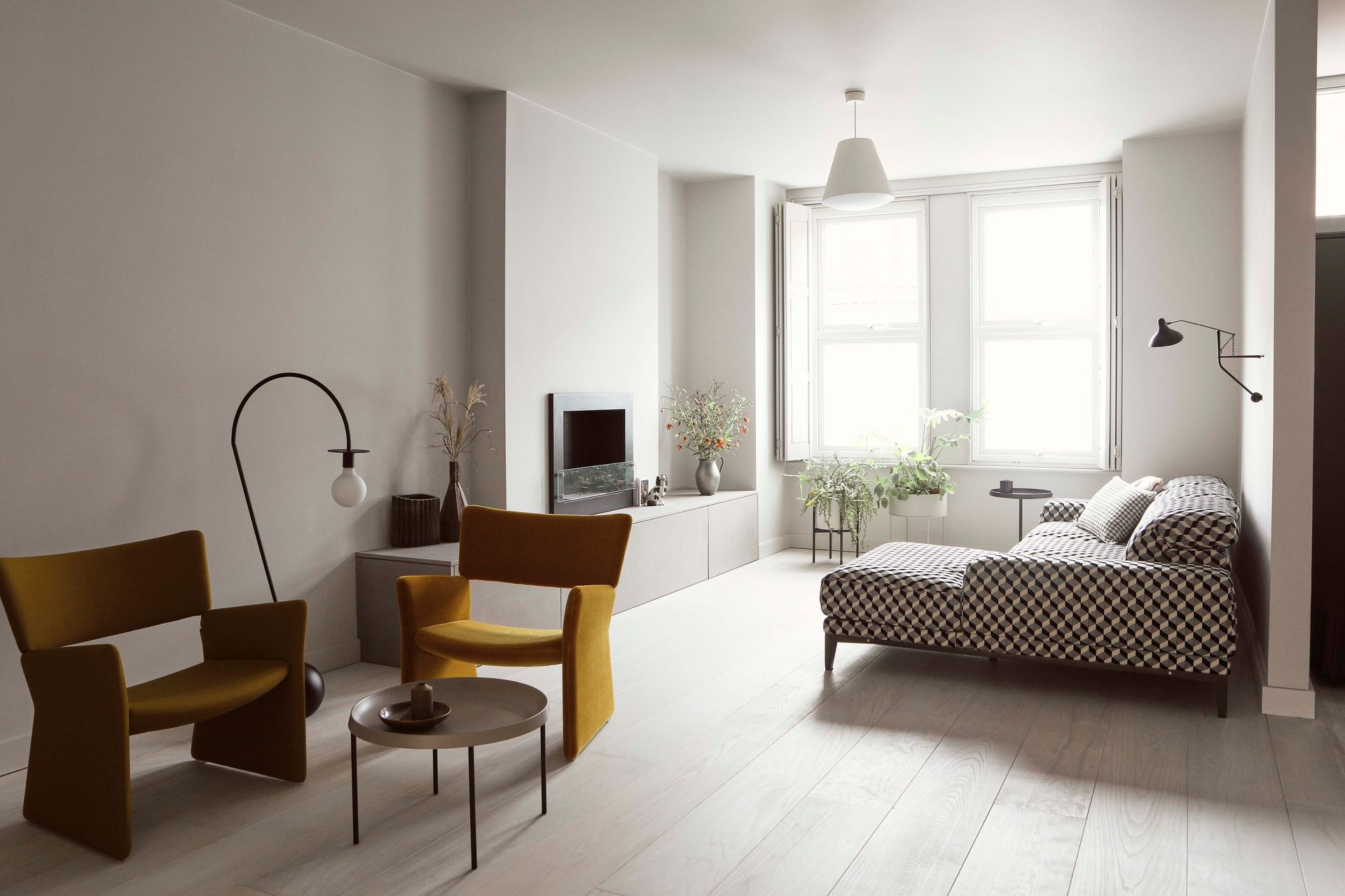 Bright minimalist living room with white walls, wood floor, tan chairs, and a low bed by large windows