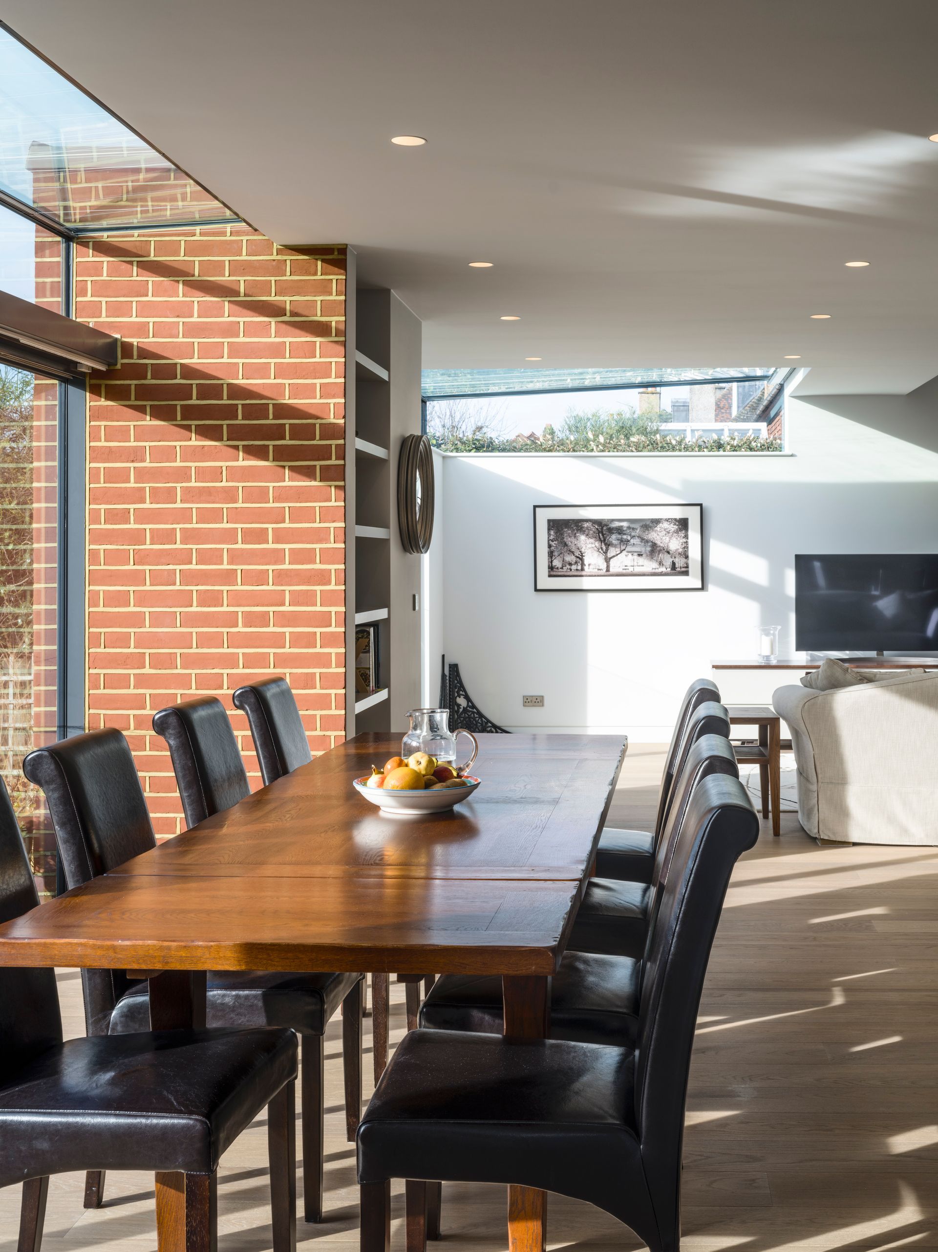 Modern dining room with a long wooden table, black chairs, and a bright living area beyond