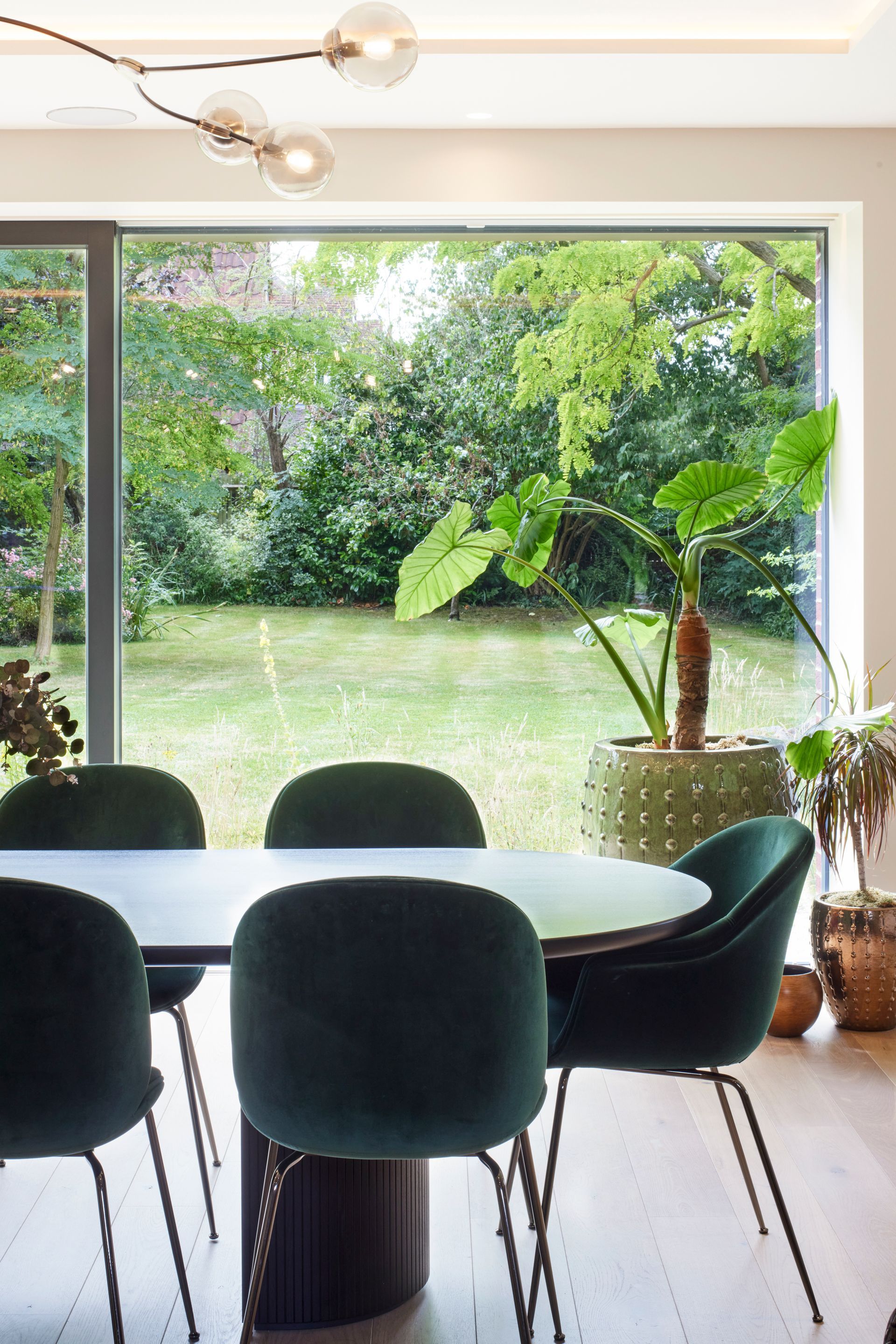 Dining table and black chairs in a bright room overlooking a lush green garden through a large window.