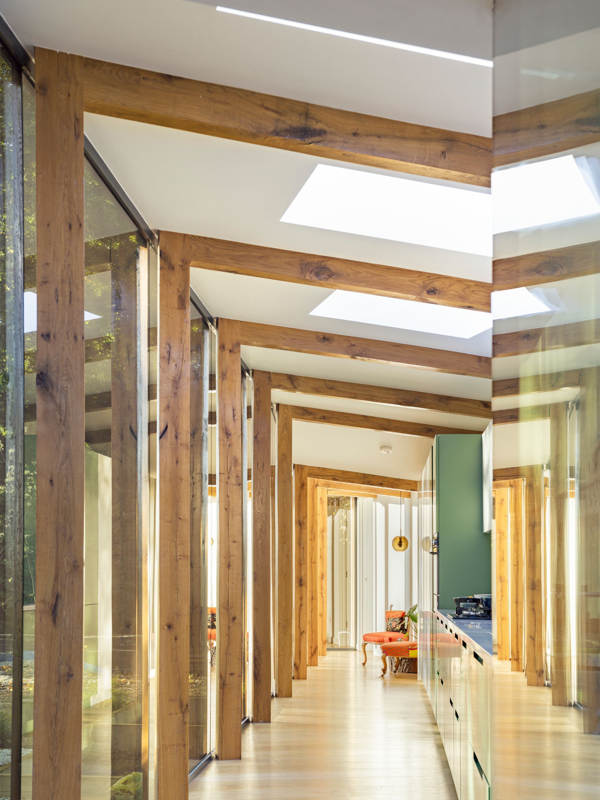 Wood-and-glass hallway with repeating frames and skylights, leading to a bright room with orange chairs.