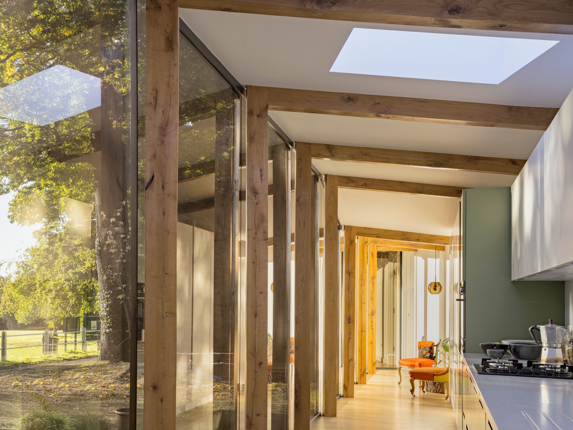 Modern corridor with wood beams, skylights, and a view of trees outside; kitchen visible on the right