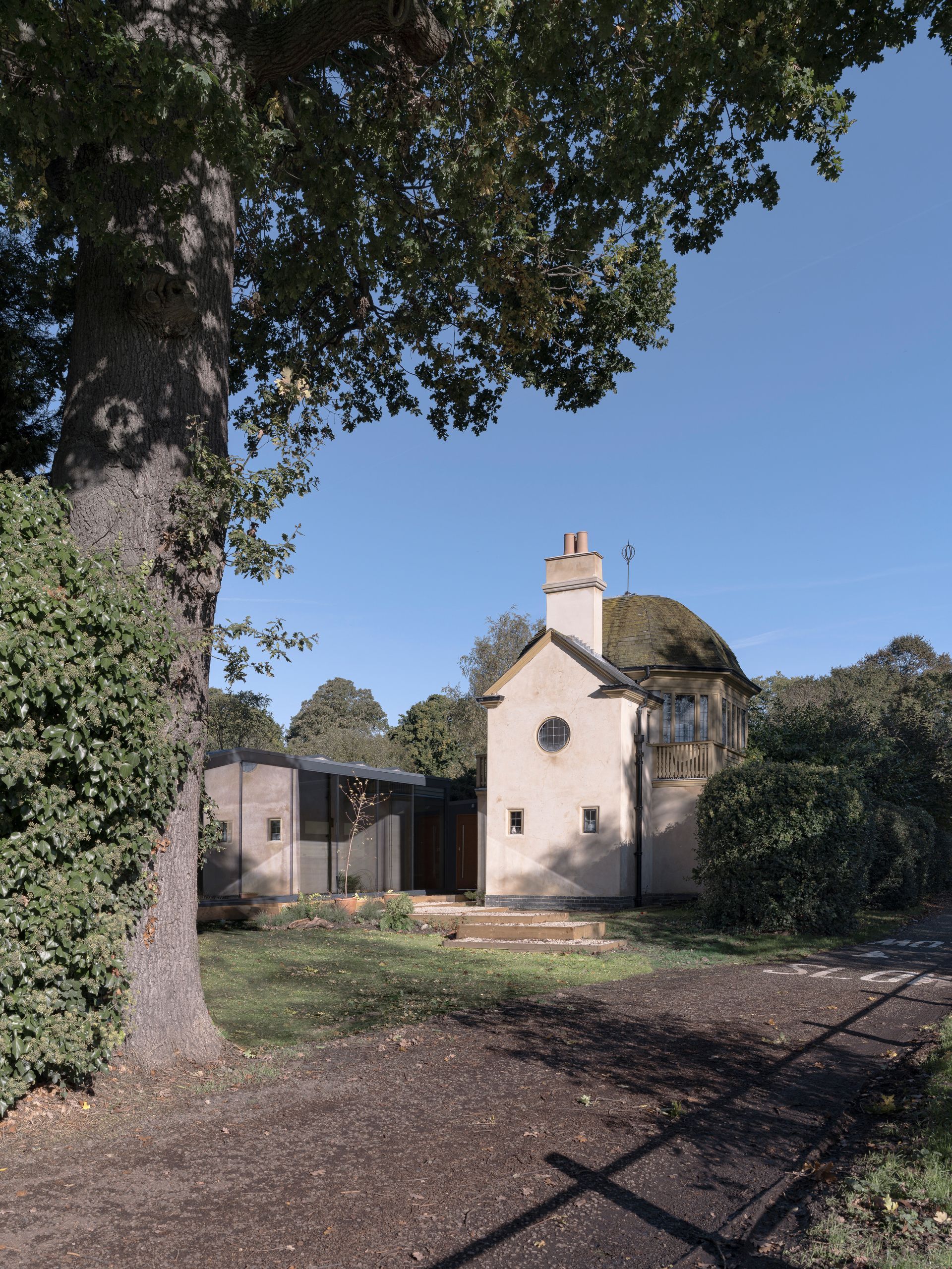 Small white house with a chimney, framed by a large tree and hedges in a sunny yard