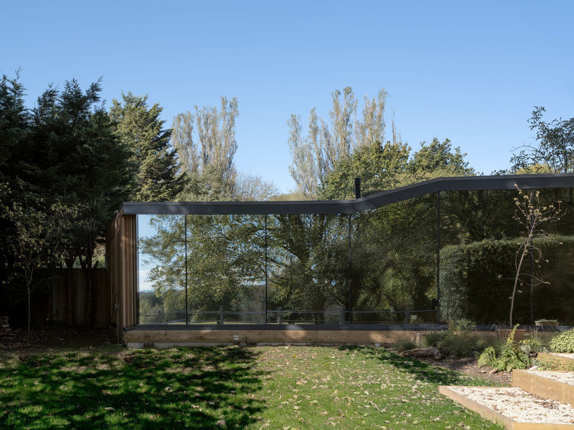 Modern house with large glass windows and a patio, set in a sunny garden with trees and grass.