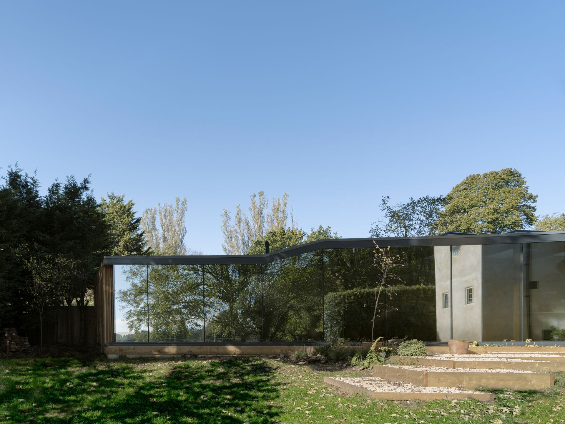 Modern backyard with glass-walled structure, patio steps, lawn, trees, and clear blue sky