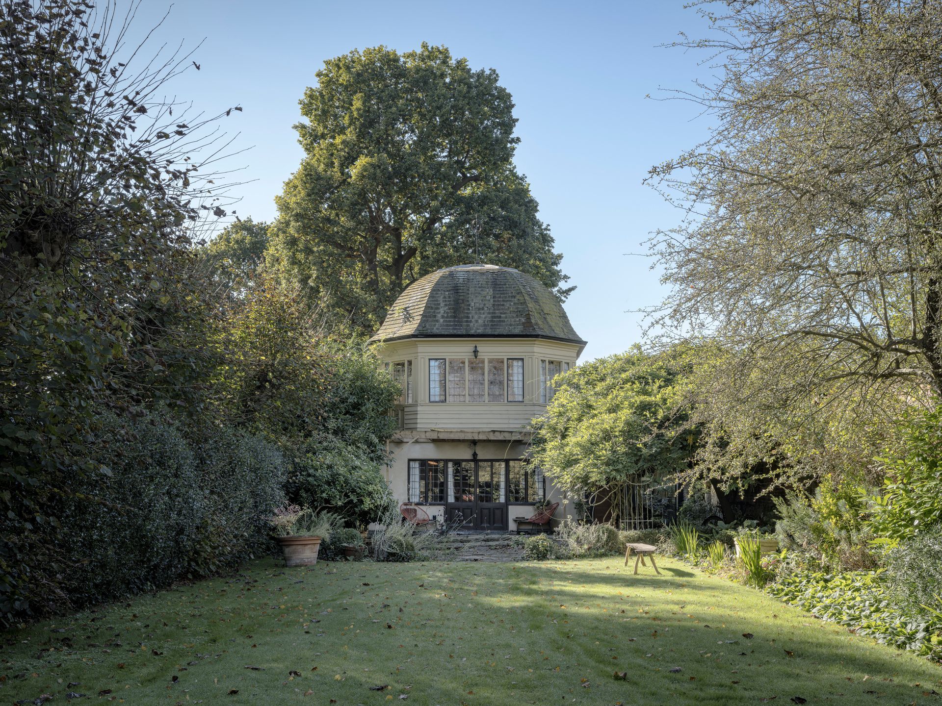Small cottage with a curved roof surrounded by a lush garden and trees under a clear blue sky