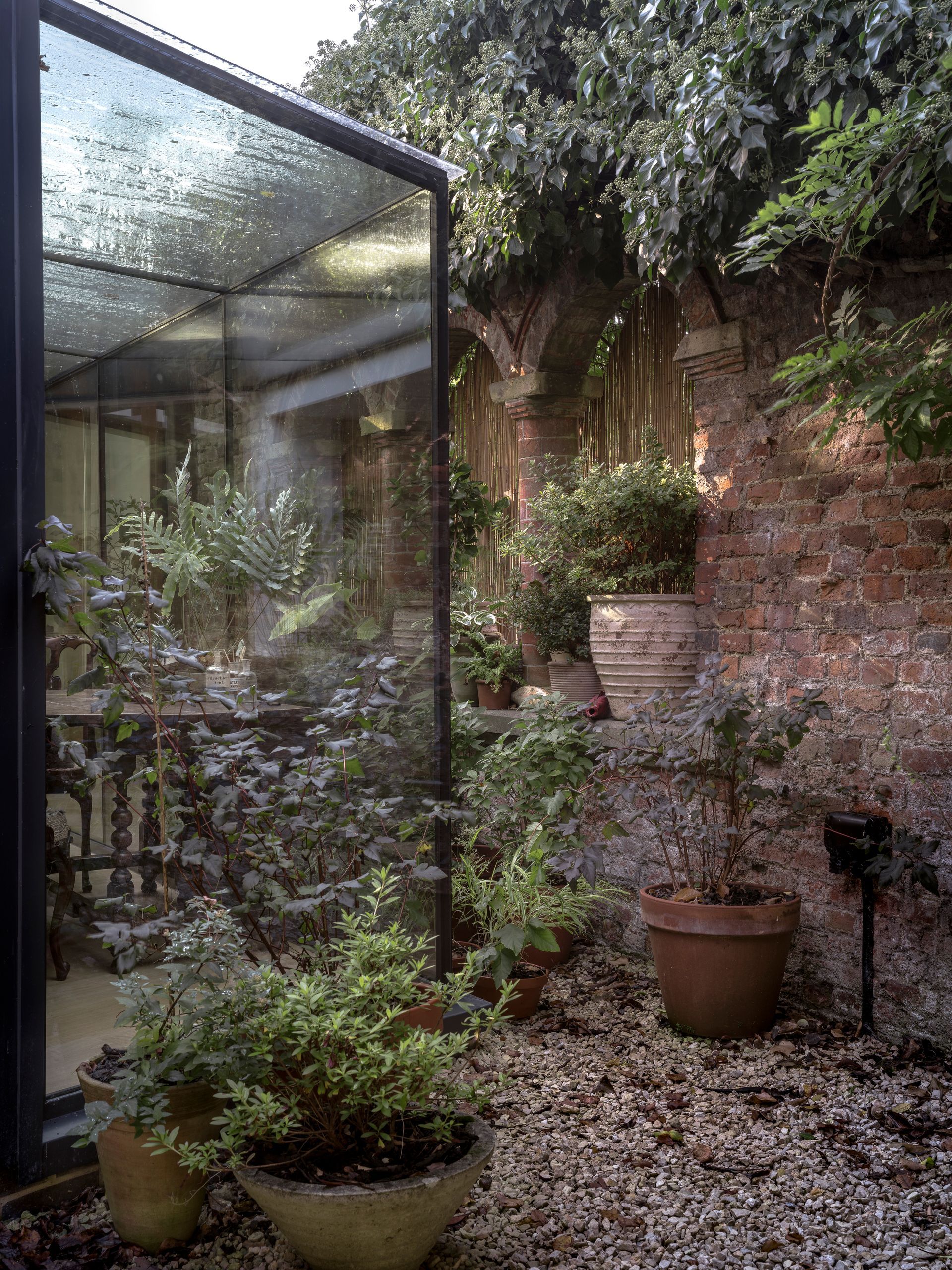 Small courtyard garden with potted plants beside a glass greenhouse and brick wall.