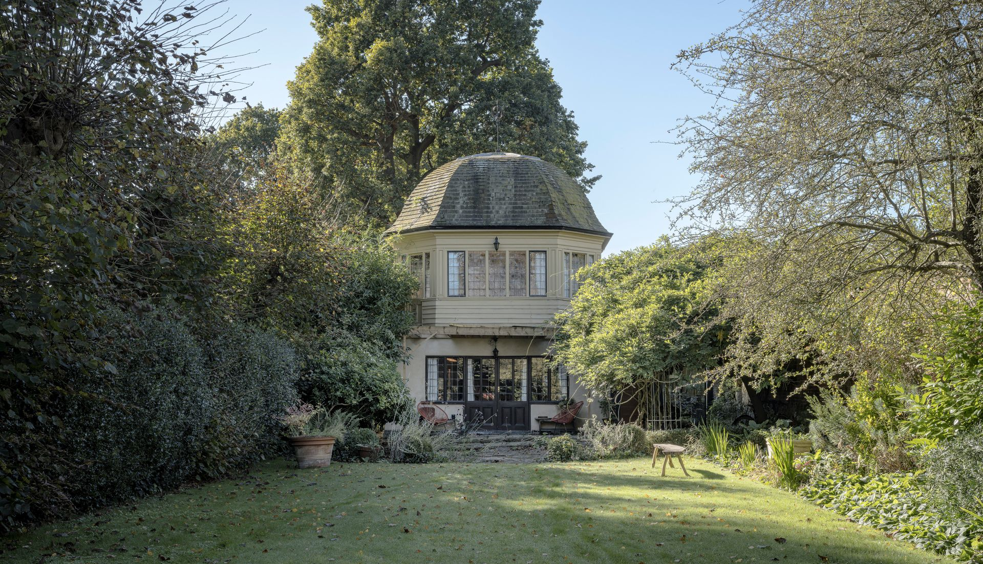 A small round tower house with a slate roof, surrounded by dense trees and a grassy garden.