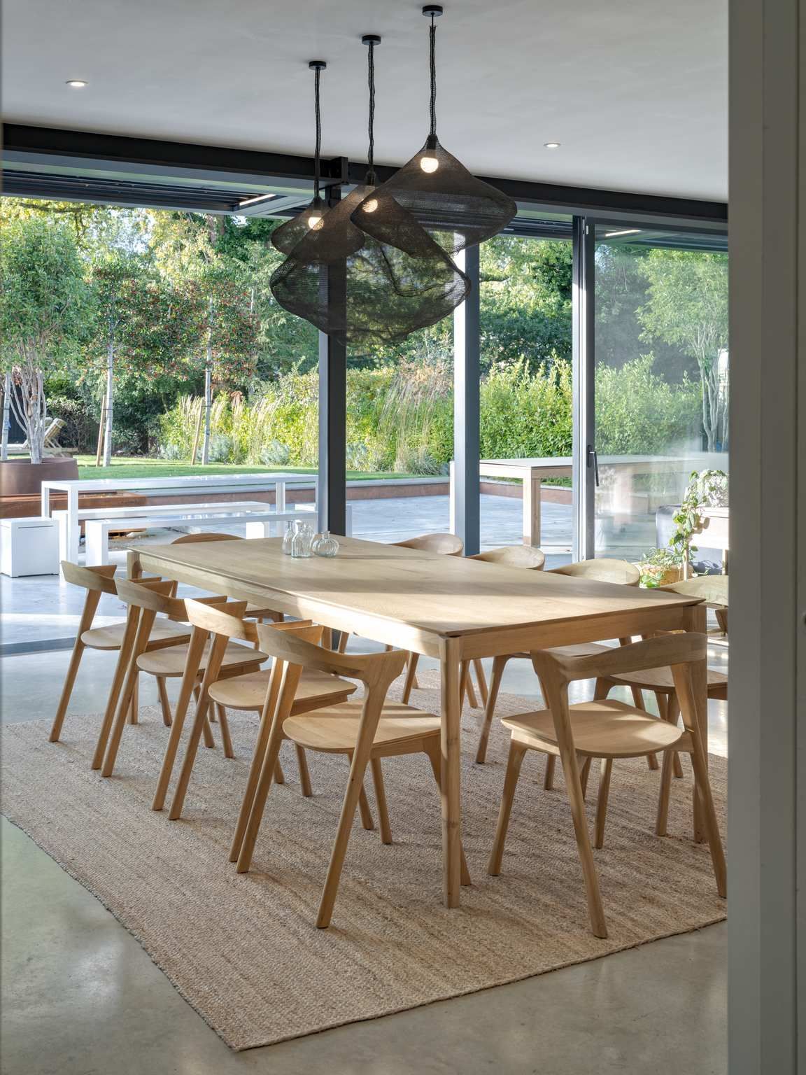 Modern dining room with a large wood table, wicker chairs, and floor-to-ceiling windows overlooking greenery