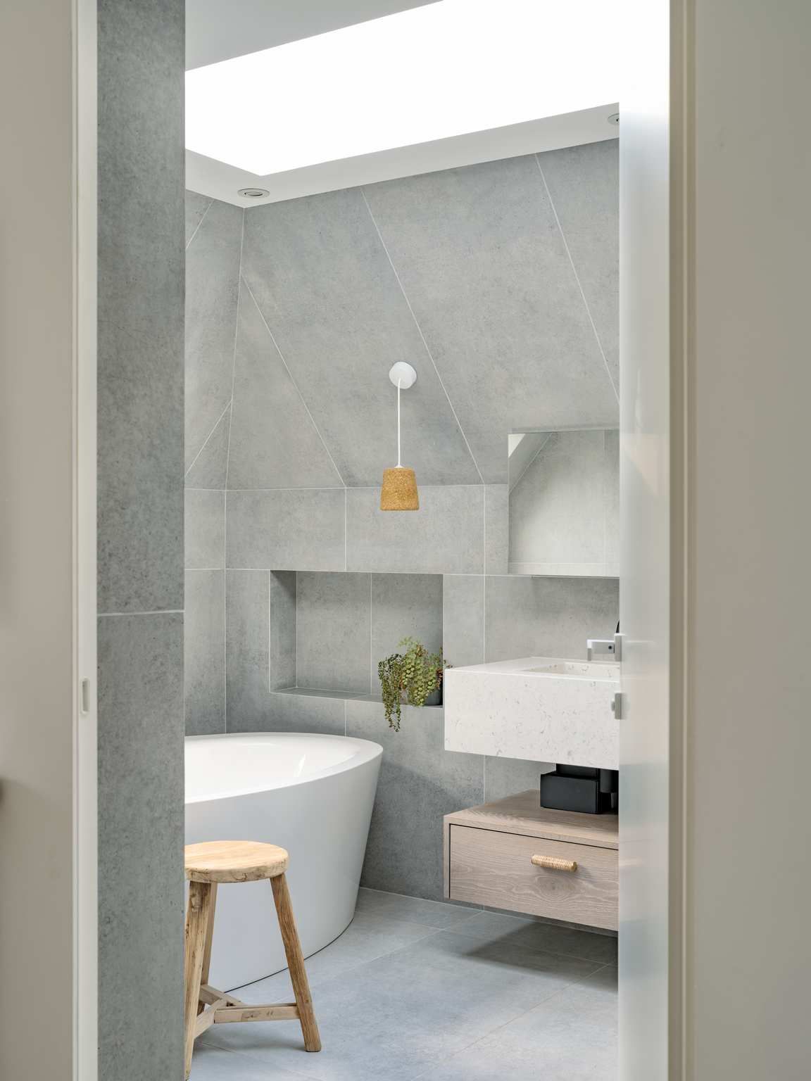 Modern gray bathroom with a freestanding tub, wall-mounted sink, and small stool under a skylight