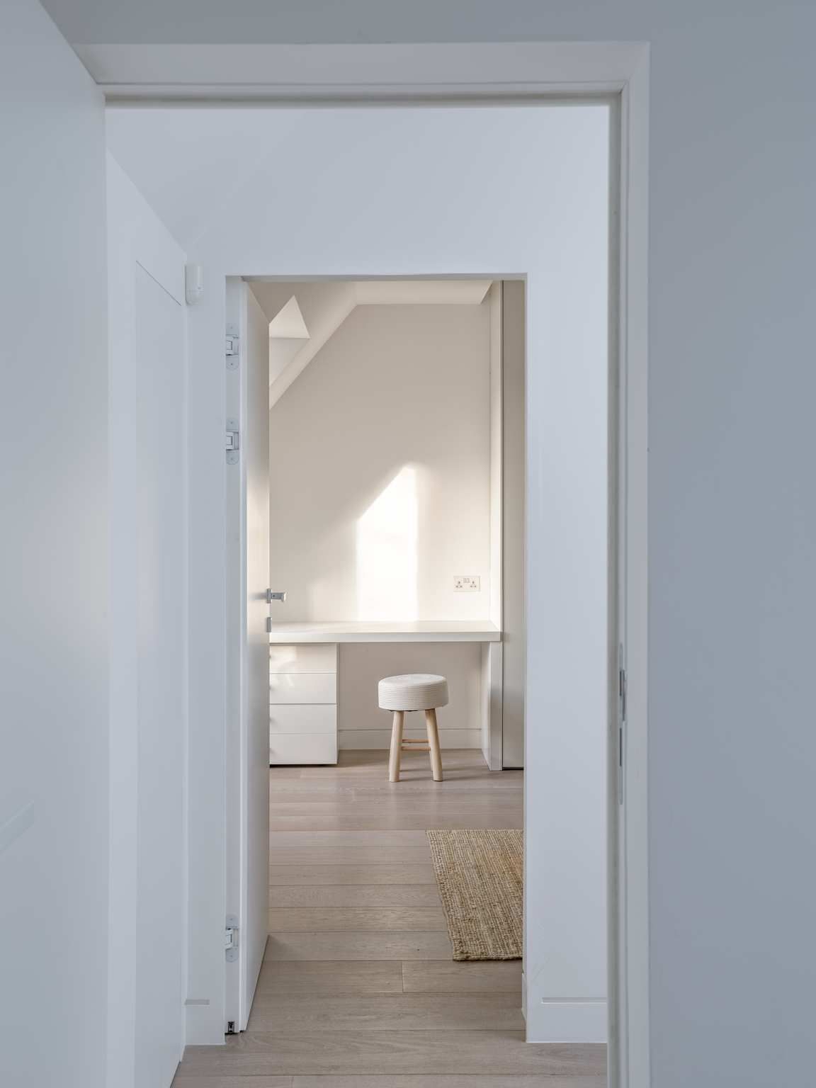 White hallway framing a bright home office with a desk, chair, and rug in a minimalist interior.