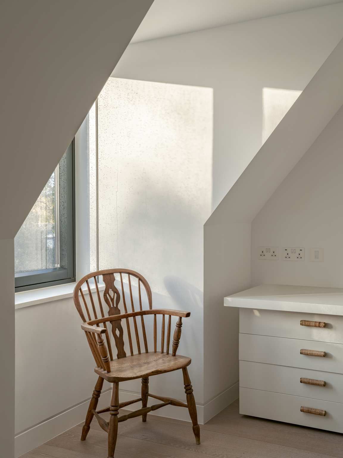 Wooden chair beside a window in a bright white attic room with a small dresser.