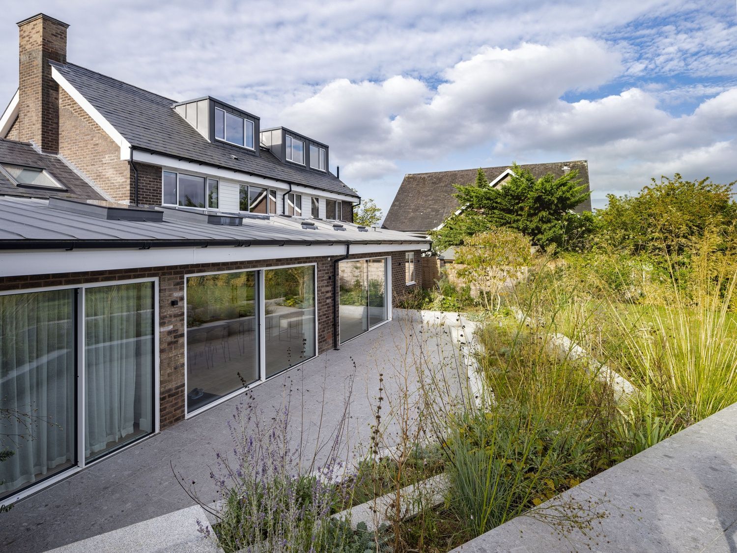 Modern house with balcony and patio beside tall overgrown grass under a cloudy sky