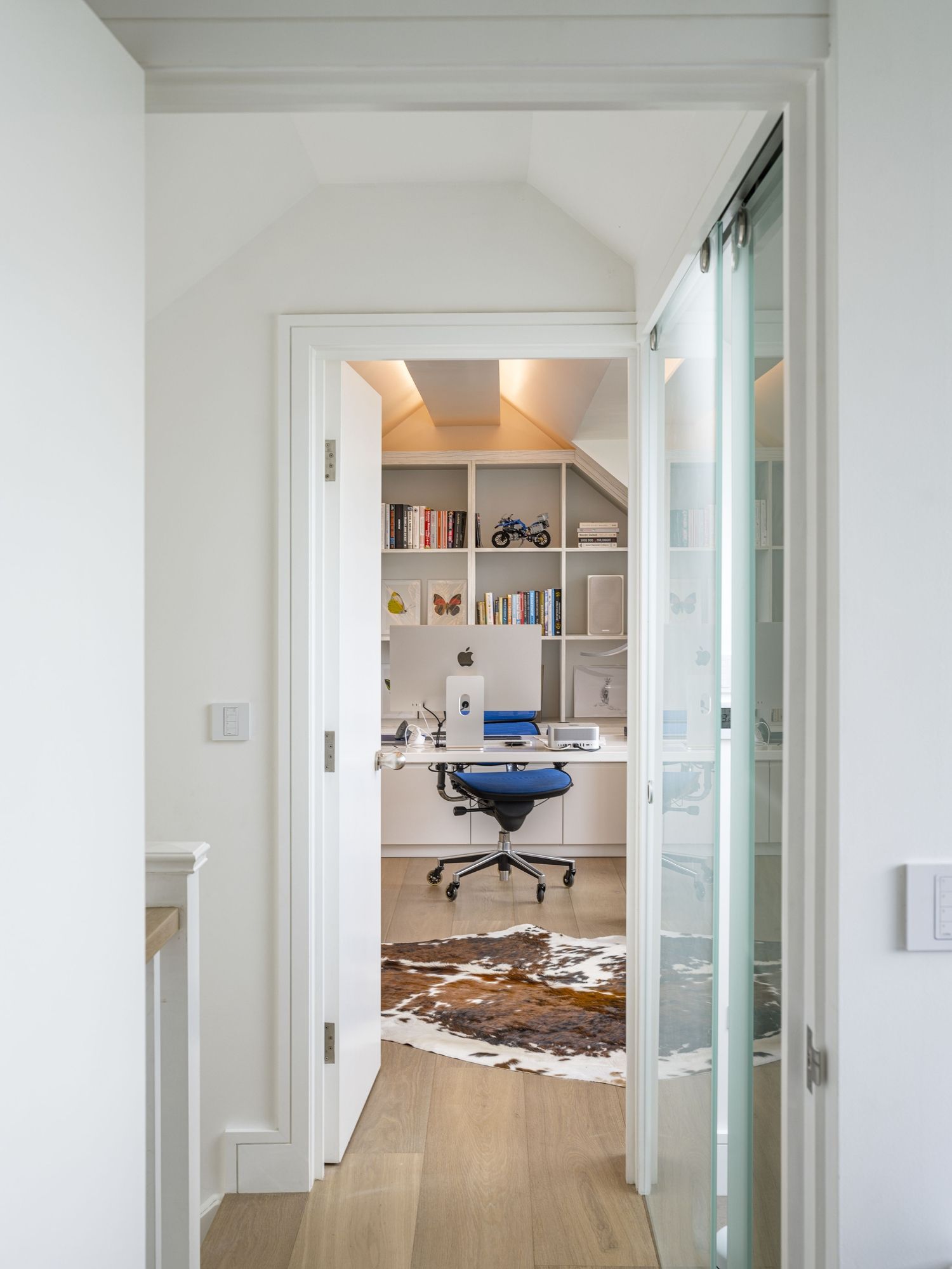 Bright home office seen through open doors, with a desk, blue chair, shelves, and a cowhide rug.