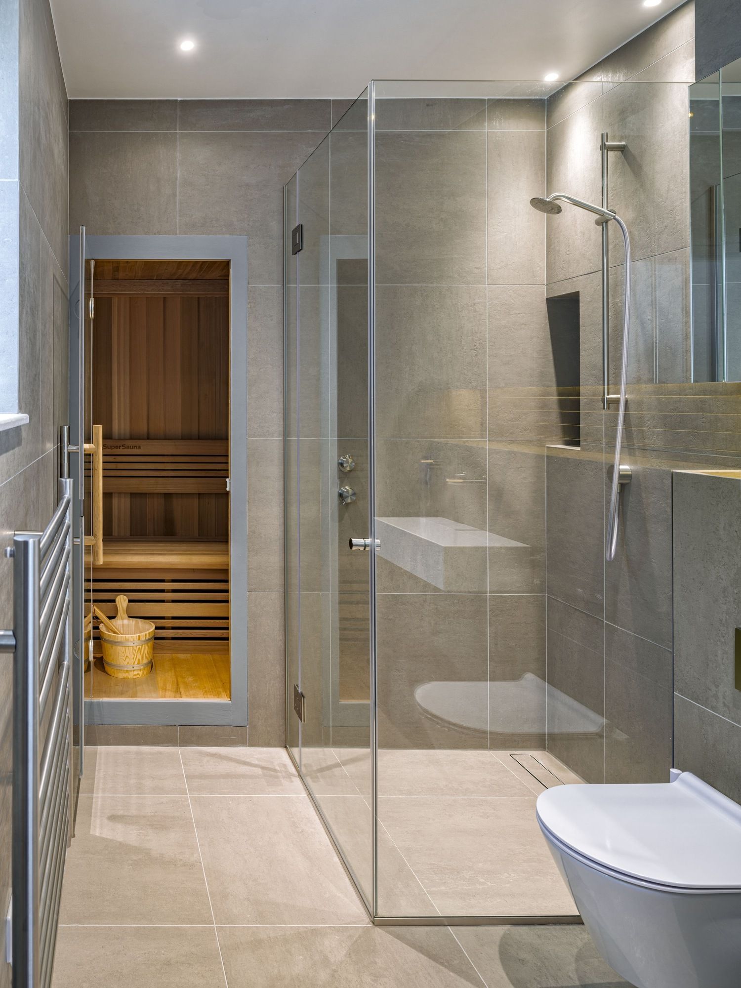 Modern bathroom with glass shower, beige tile, and a wooden sauna room in the background