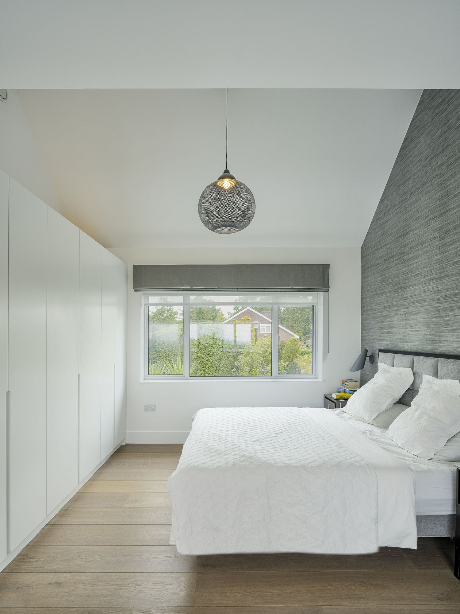 Bright modern bedroom with white bed, built-in closet, gray accent wall, and large window overlooking greenery