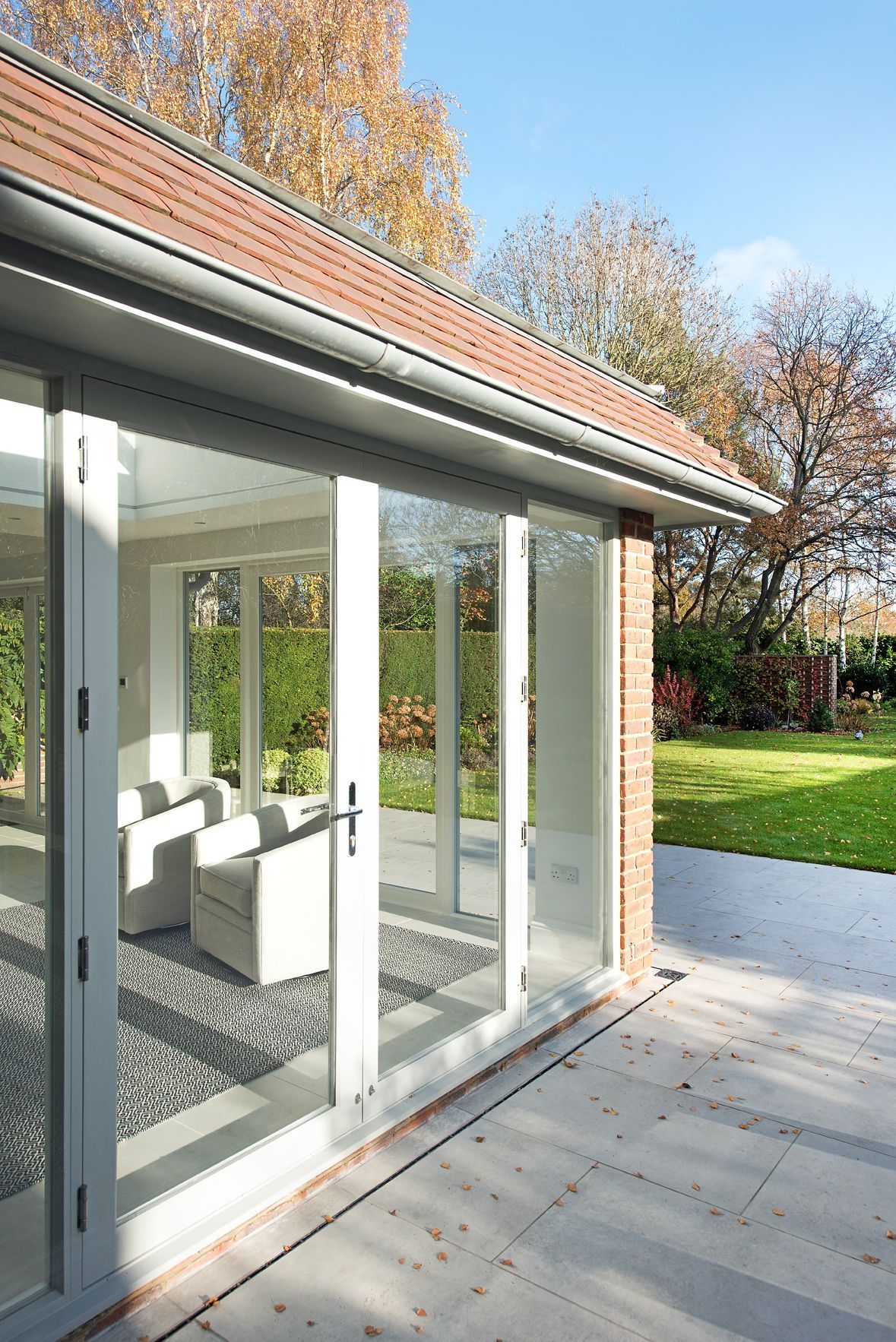 Modern glass patio room beside a brick house, opening to a sunny garden and lawn