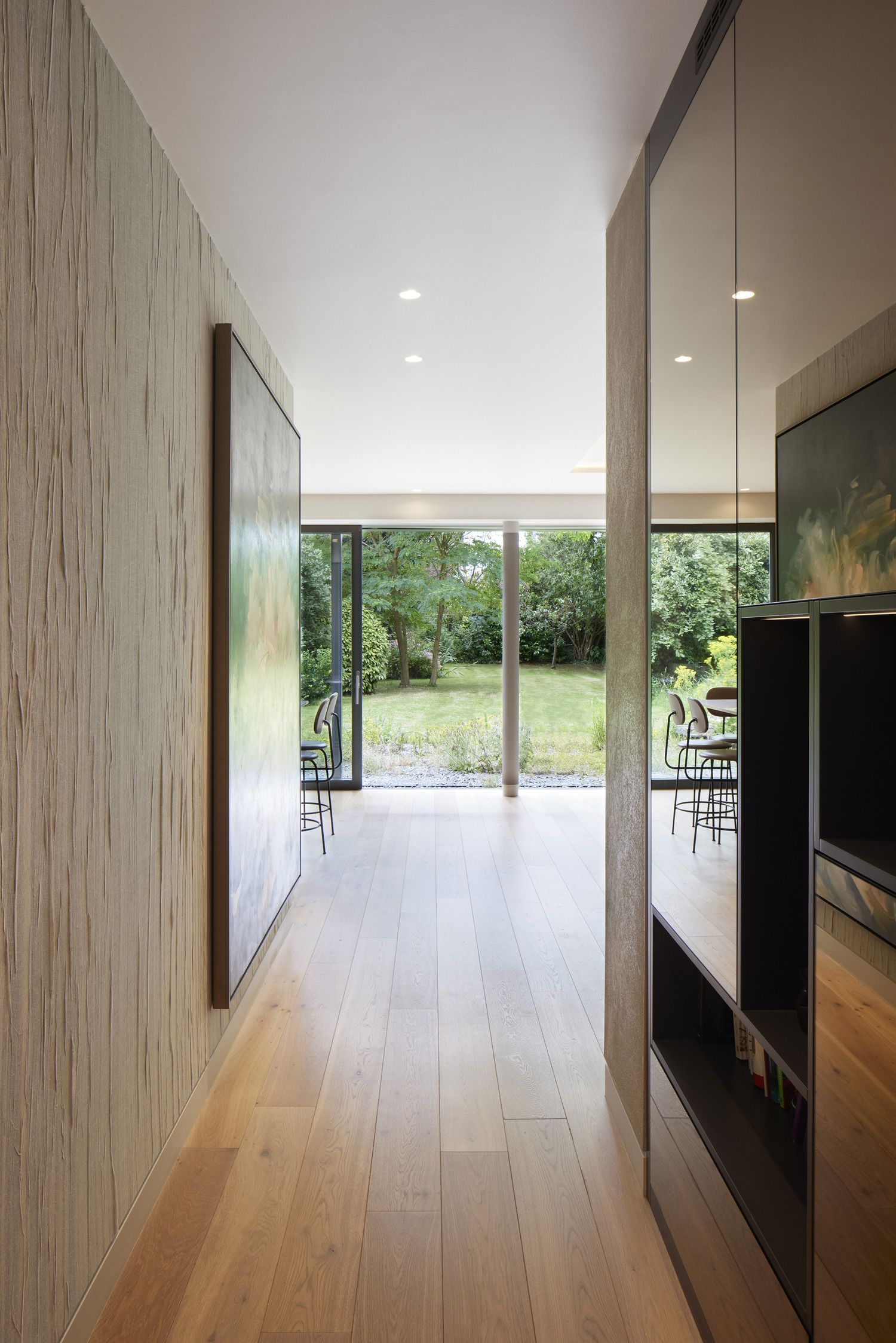 Modern hallway with wood floors, white walls, and open glass doors to a green backyard.