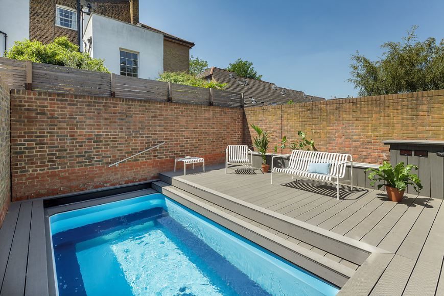 Backyard deck with a small blue pool, lounge chairs, and a brick wall in sunlight