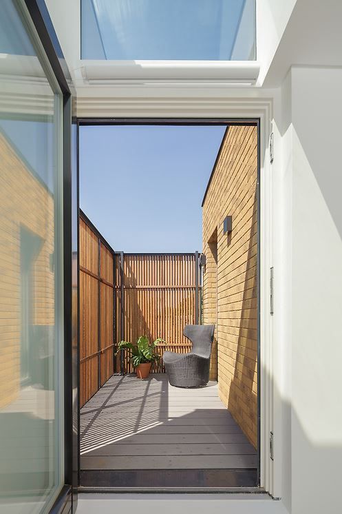 Narrow rooftop patio with wooden deck, potted plants, and gray chair beside brick walls under a skylight.