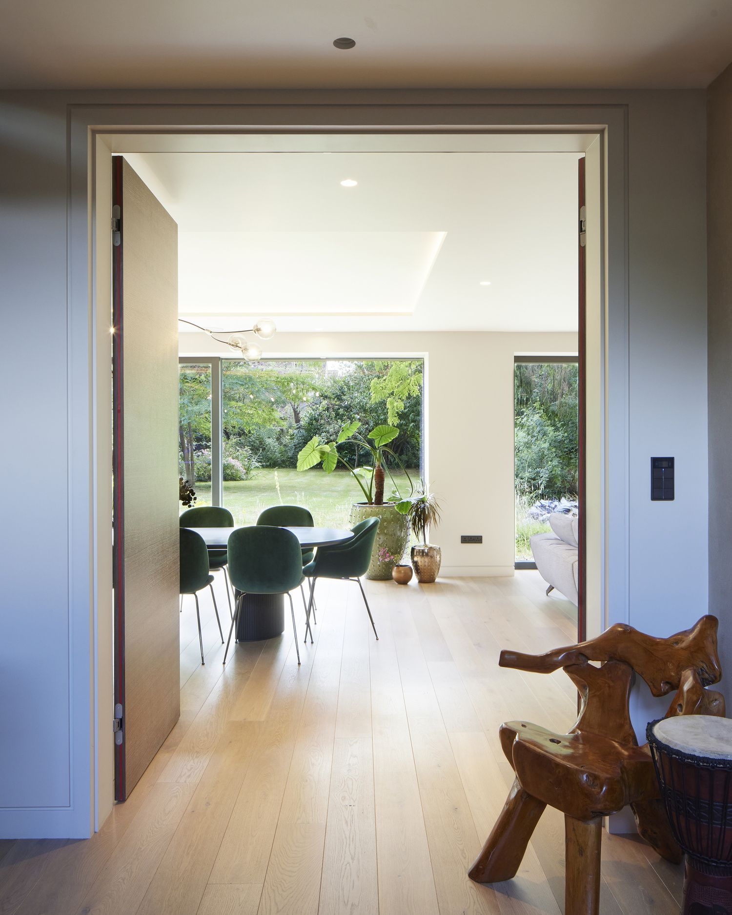 Open doorway to a bright dining room with green chairs and garden views; wooden sculpture in foreground