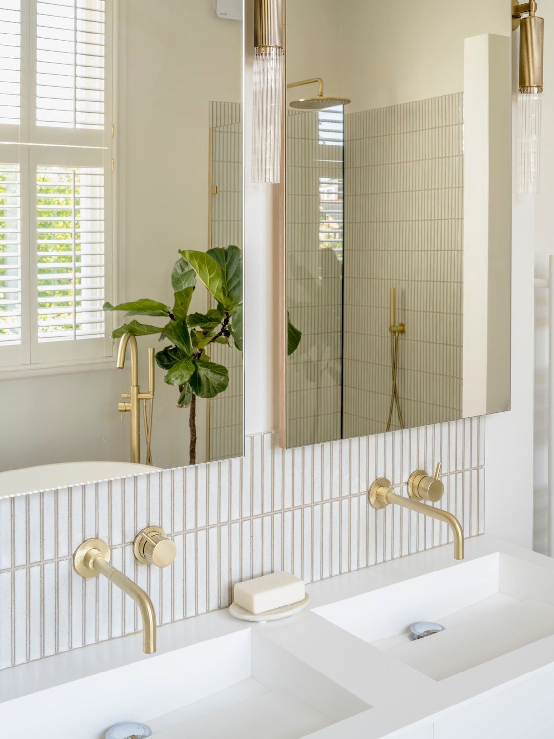 Bright bathroom vanity with double sinks, brass faucets, mirror, and a leafy plant by a window.