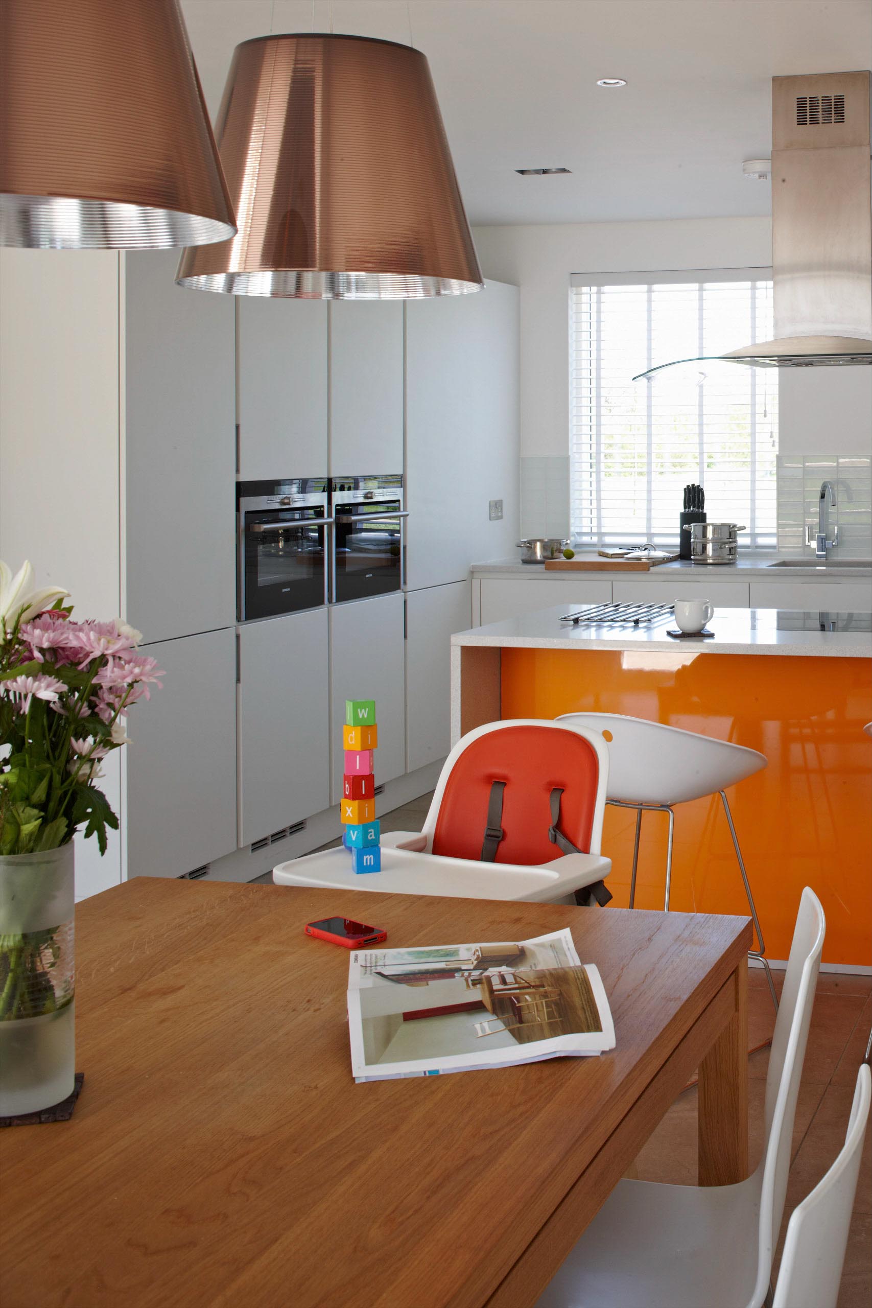 Modern kitchen and dining area with orange cabinets, wood table, high chair, and hanging copper lamps