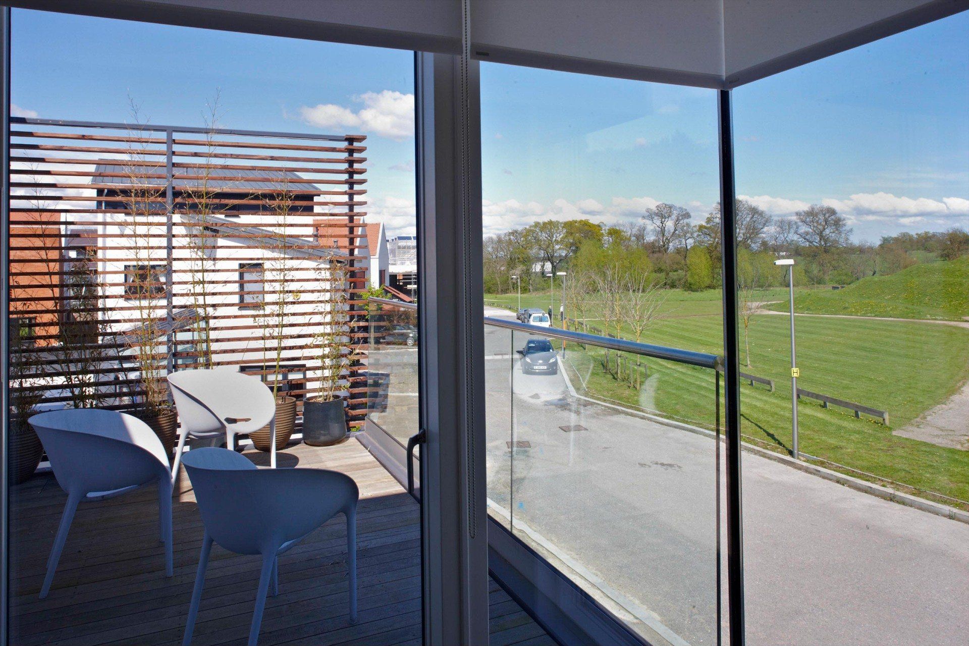 Balcony with white chairs overlooking a green field and road, seen through glass walls