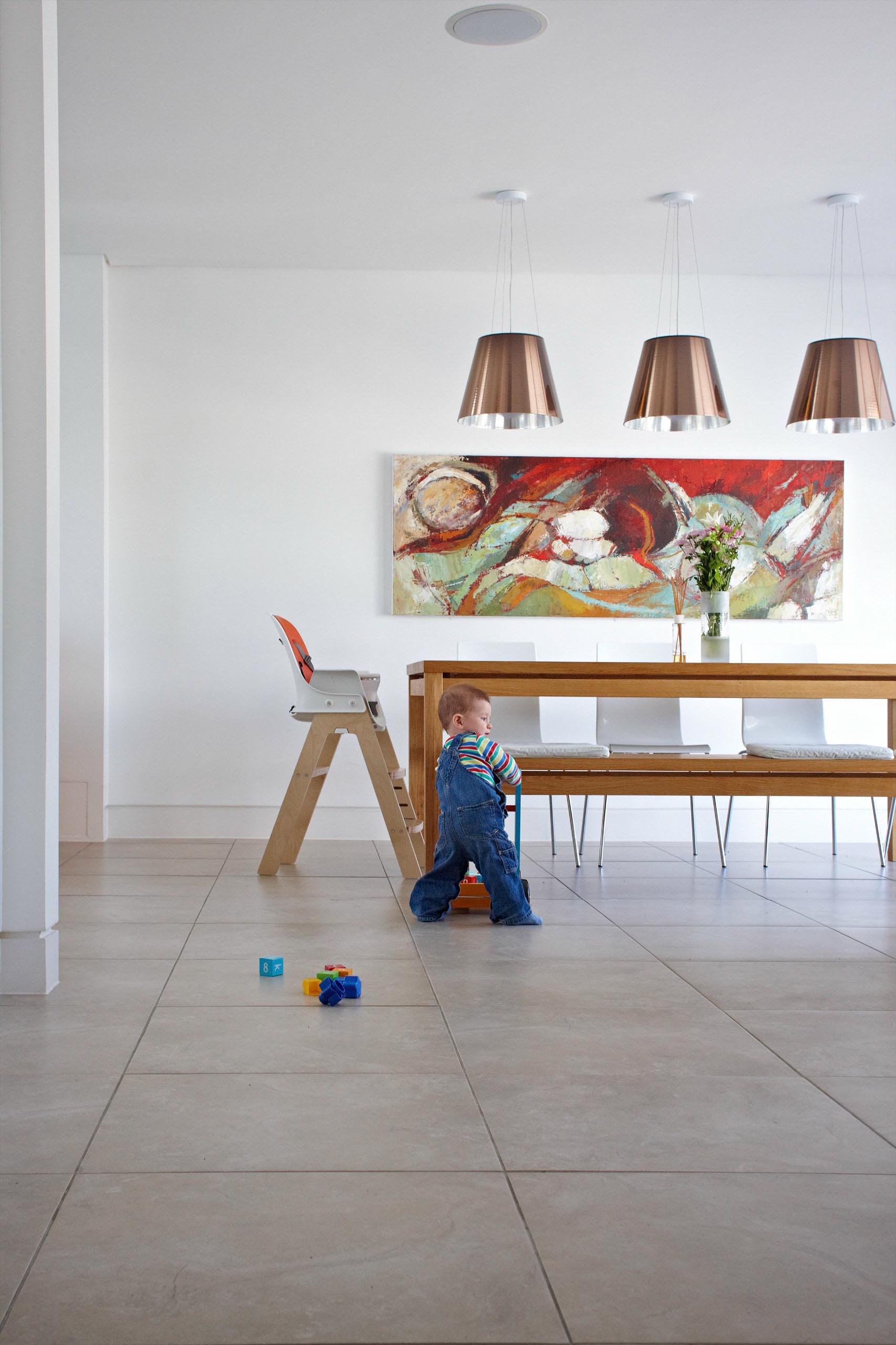 Toddler on a tiled floor in a bright dining room, near a wooden table and colorful wall art.